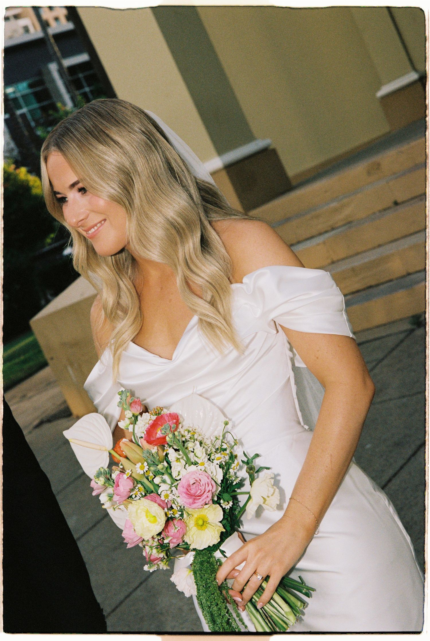 Smiling blonde bride in off-shoulder white satin gown holds colorful pink and yellow floral bouquet.