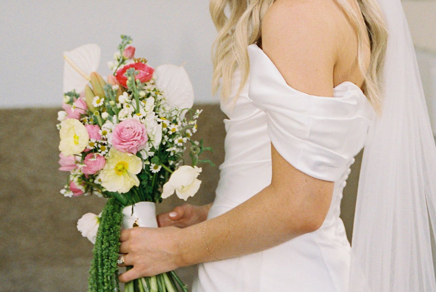 Bride in elegant off-shoulder white satin gown holding a vibrant pink and yellow floral bouquet.