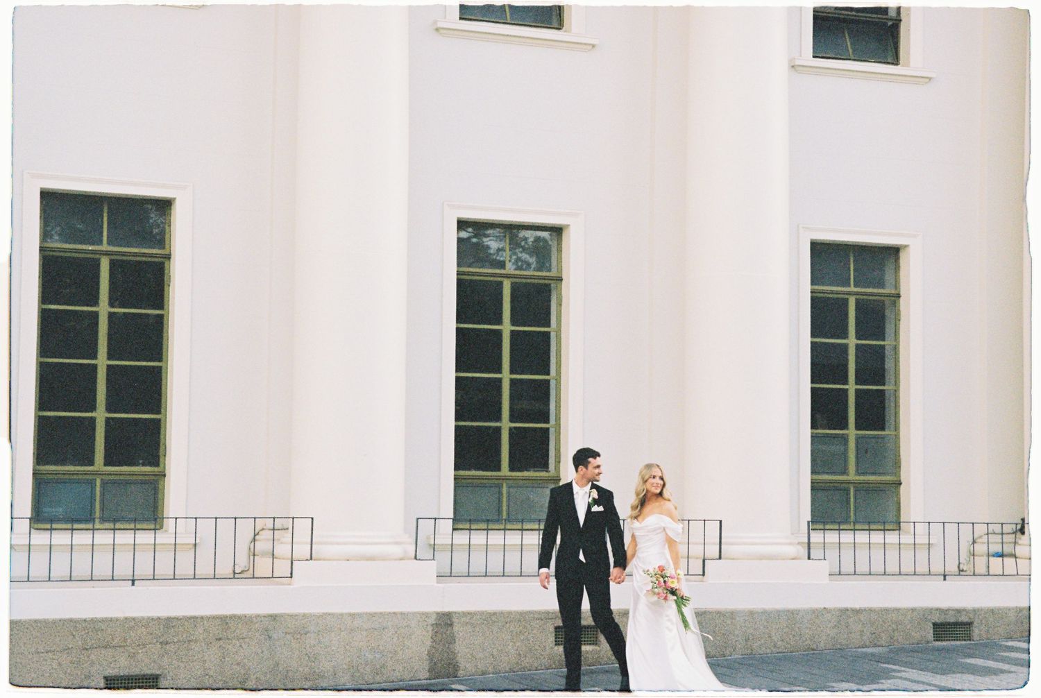 Bride and groom walk hand in hand along a white neoclassical building on their wedding day.