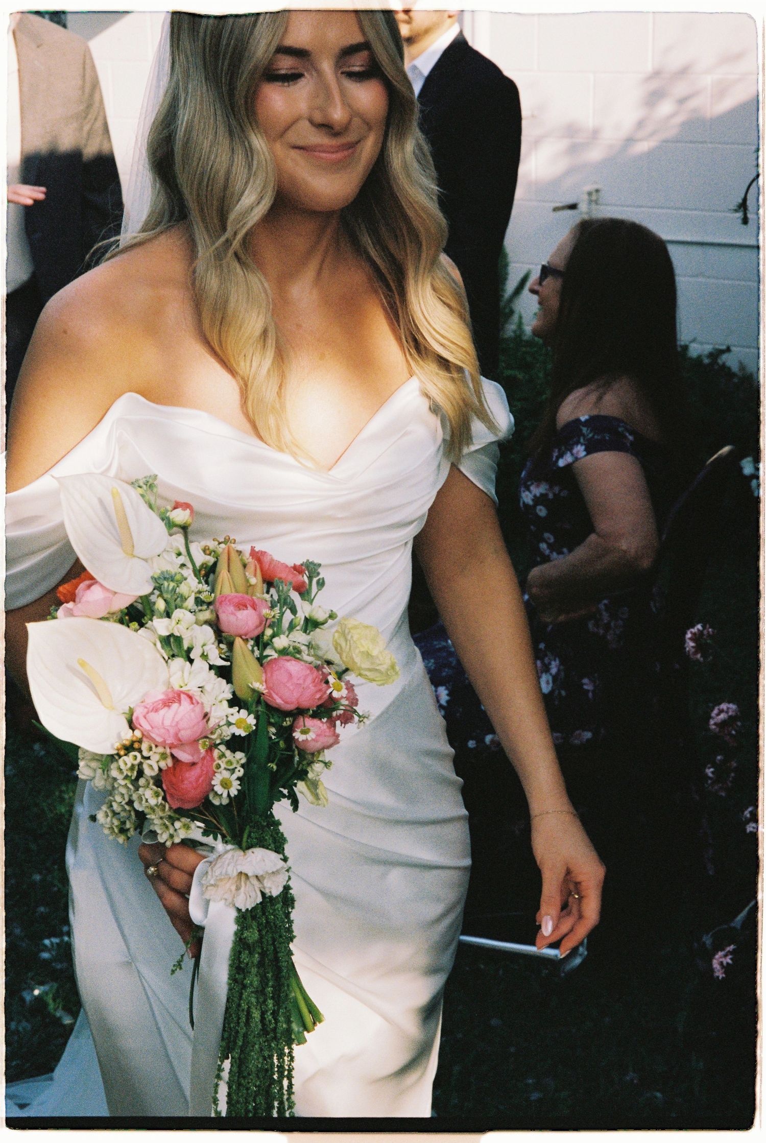 Smiling bride in white off-shoulder gown holding pink and white floral bouquet at outdoor wedding ceremony.