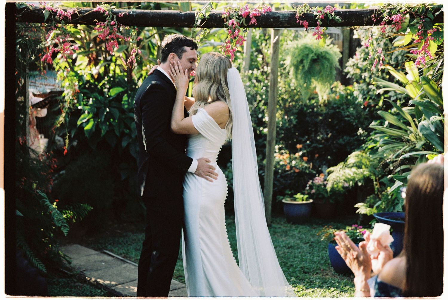 Bride and groom share first kiss at lush garden wedding ceremony surrounded by tropical greenery and flowers.