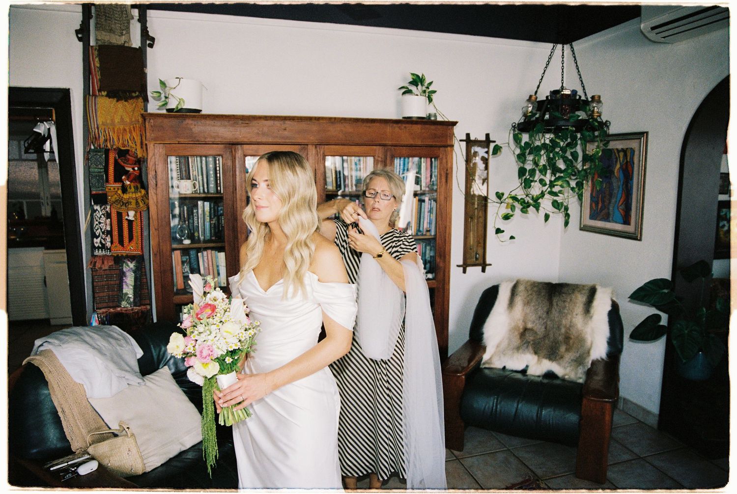 Bride holding bouquet while older woman helps adjust white wedding dress from behind.