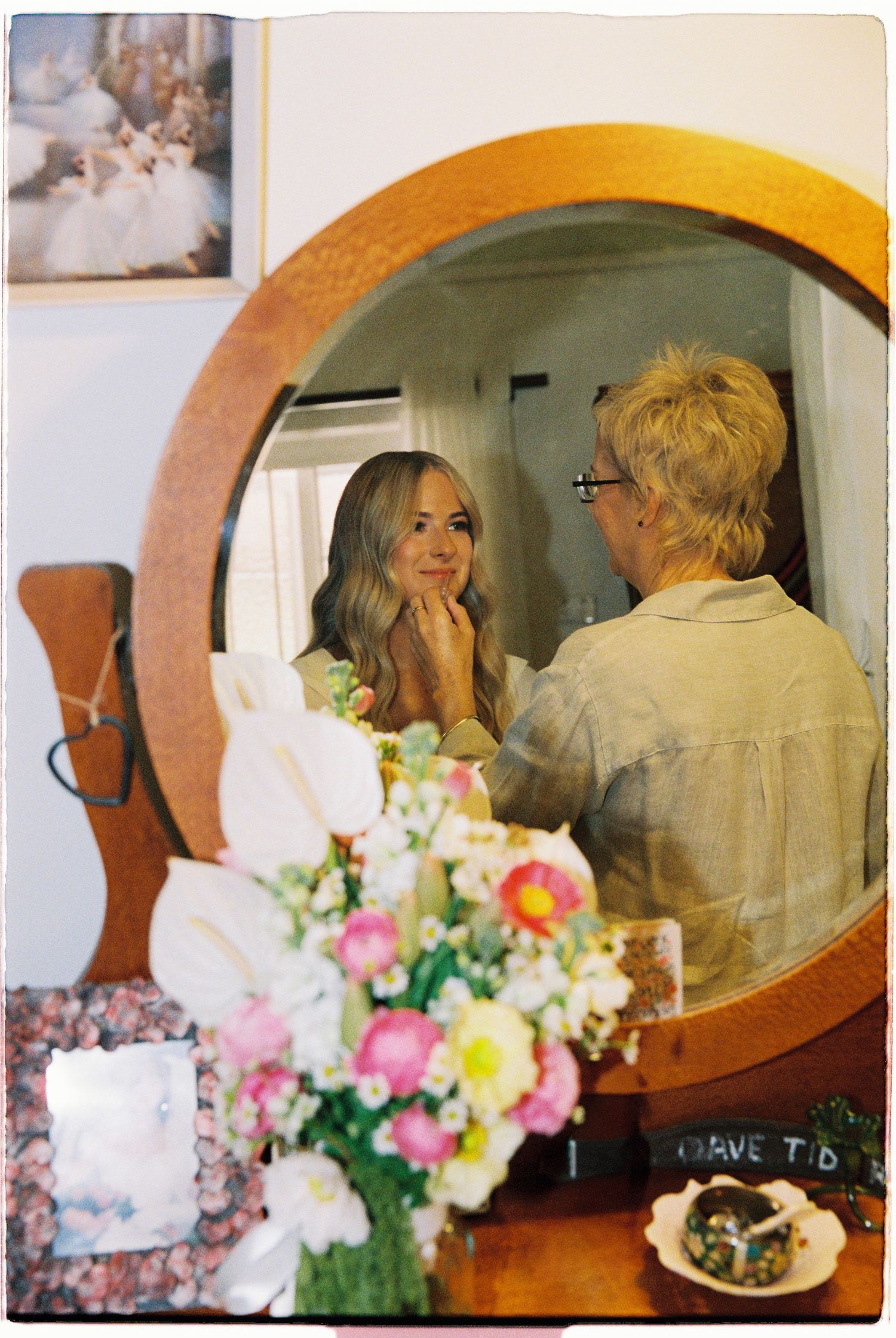 Blonde bride smiling as makeup artist applies finishing touches reflected in vintage round mirror.