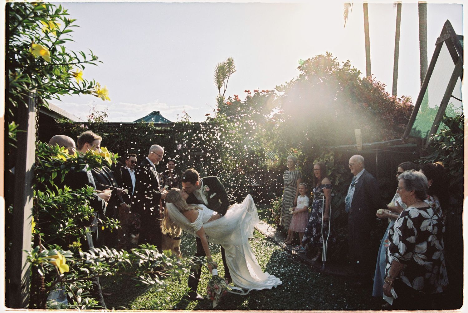 Bride and groom celebrated with confetti shower by guests in sunny outdoor garden wedding.