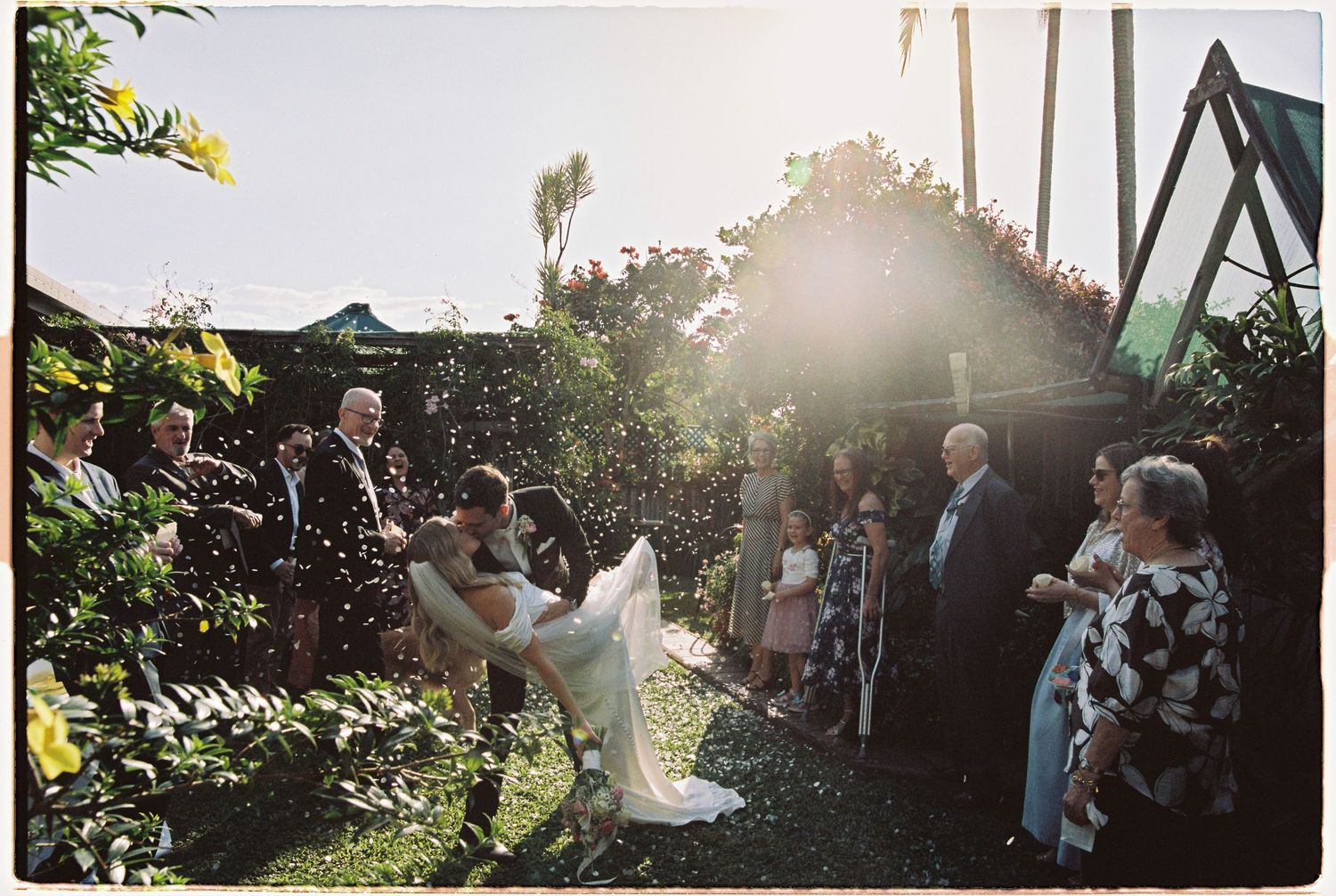 Bride and groom celebrated with confetti toss by guests during outdoor backyard wedding ceremony in golden sunlight.
