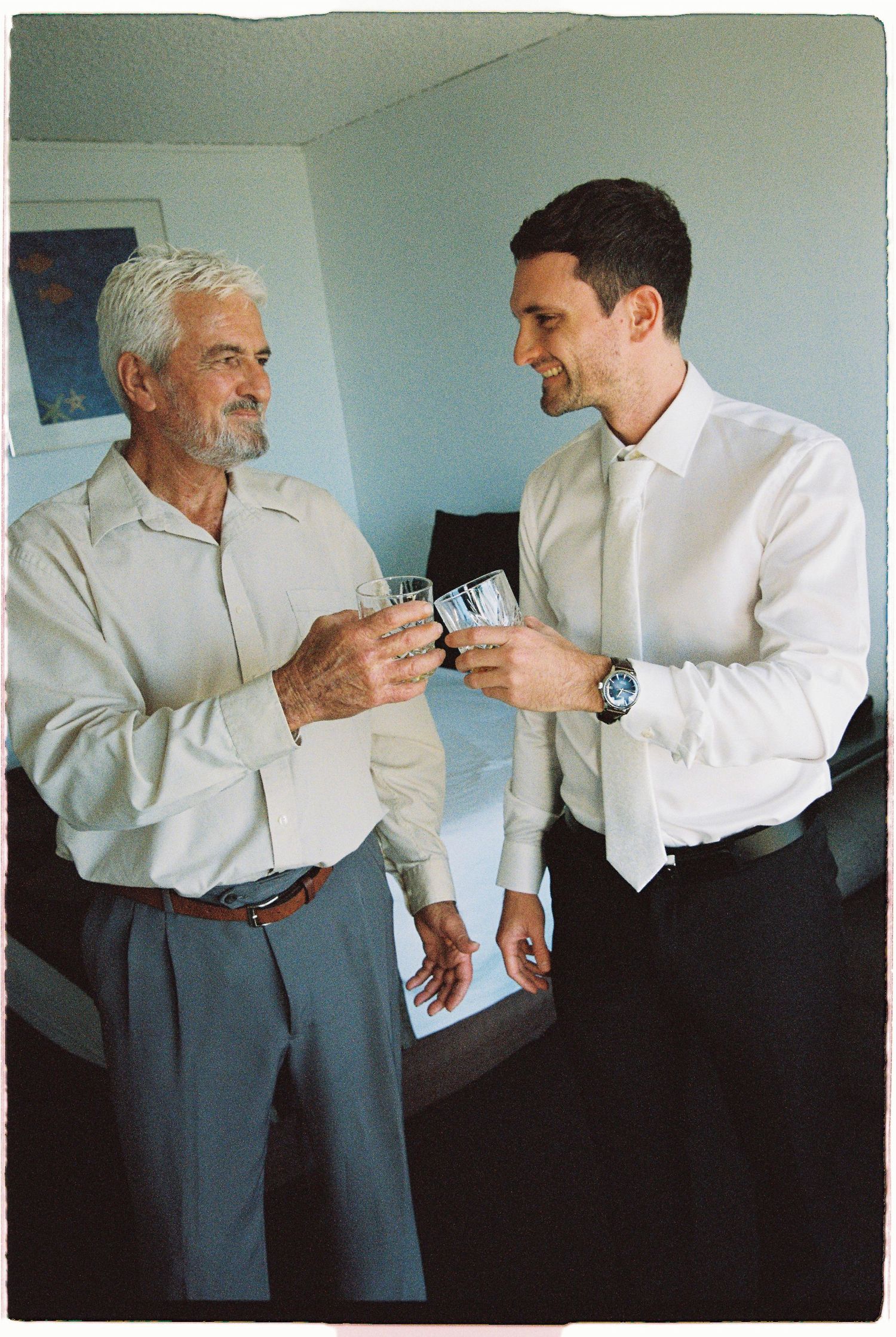 Two men in white dress shirts smiling and toasting with glasses in a hotel room.