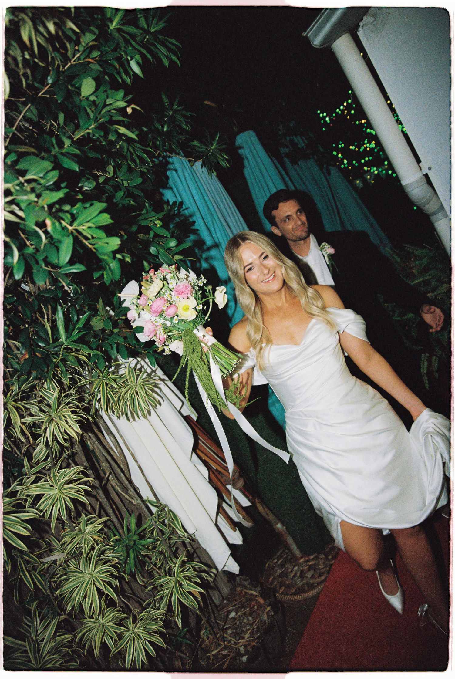 A smiling blonde bride in a white mini dress holds a pink and white bouquet walking with her groom at night.