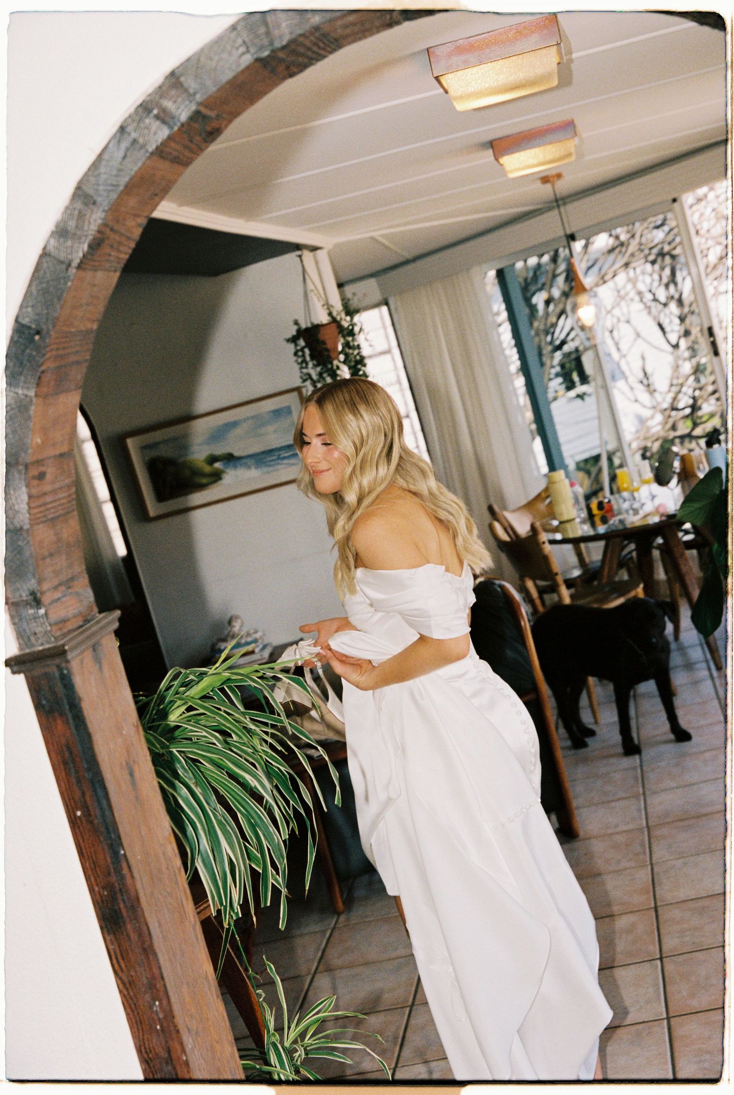Blonde bride in white off-shoulder wedding dress standing in rustic archway doorway.