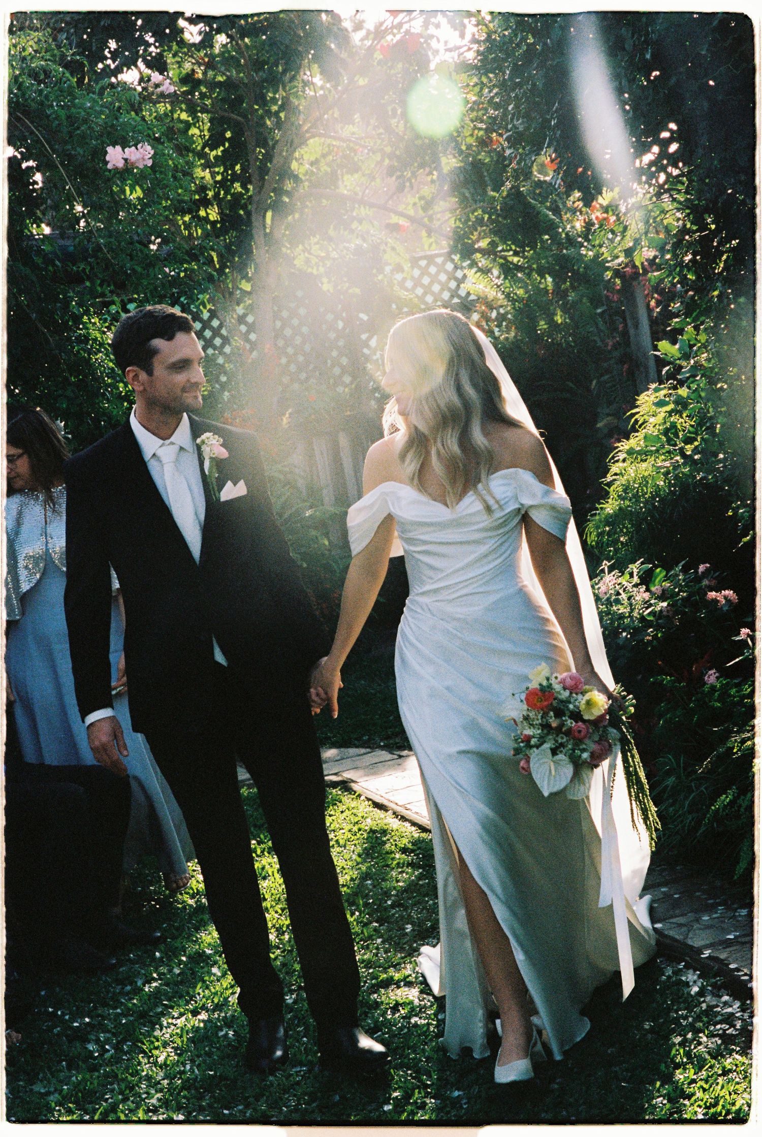 Newlyweds walk hand-in-hand through sunlit garden path after outdoor wedding ceremony.