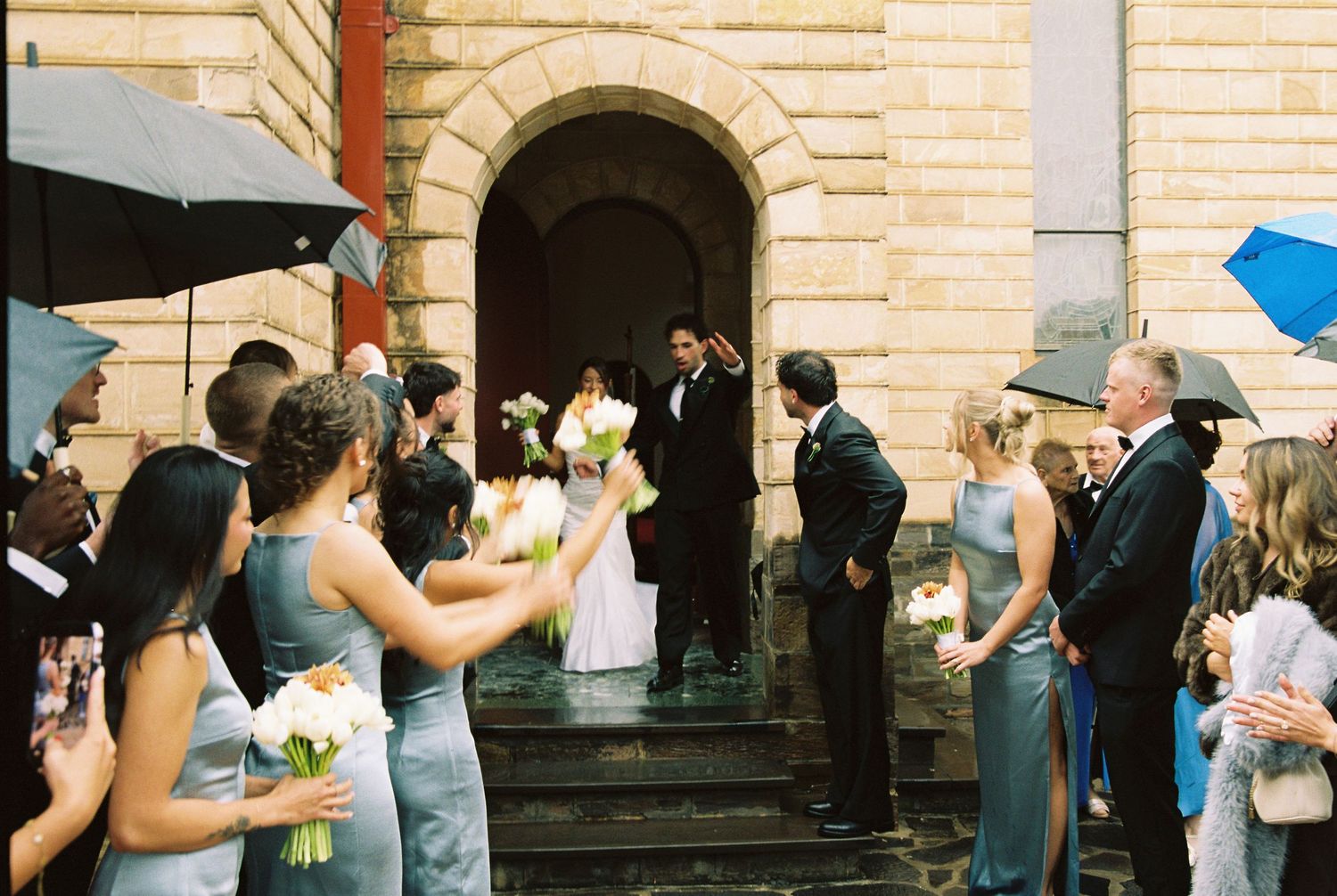 Wedding ceremony on church steps with bride, groom, bridesmaids in gray dresses, and guests holding umbrellas.