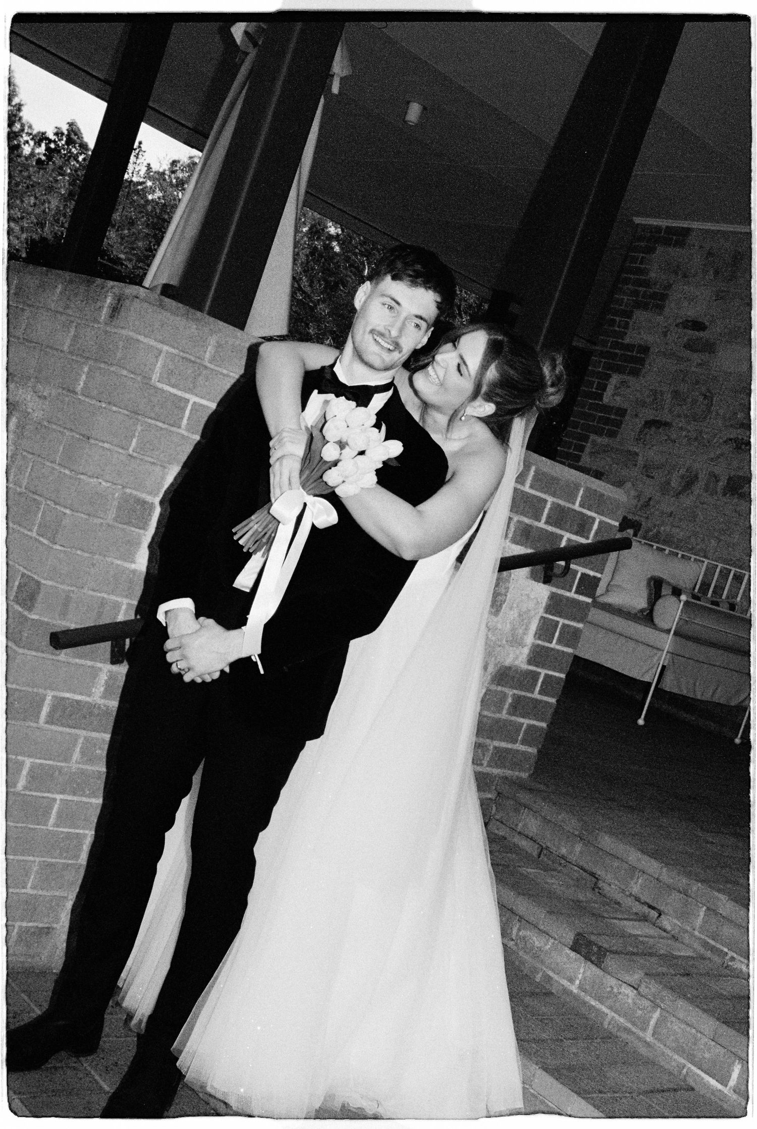 Black and white wedding photo of a smiling bride and groom embracing near a brick wall holding a bouquet.