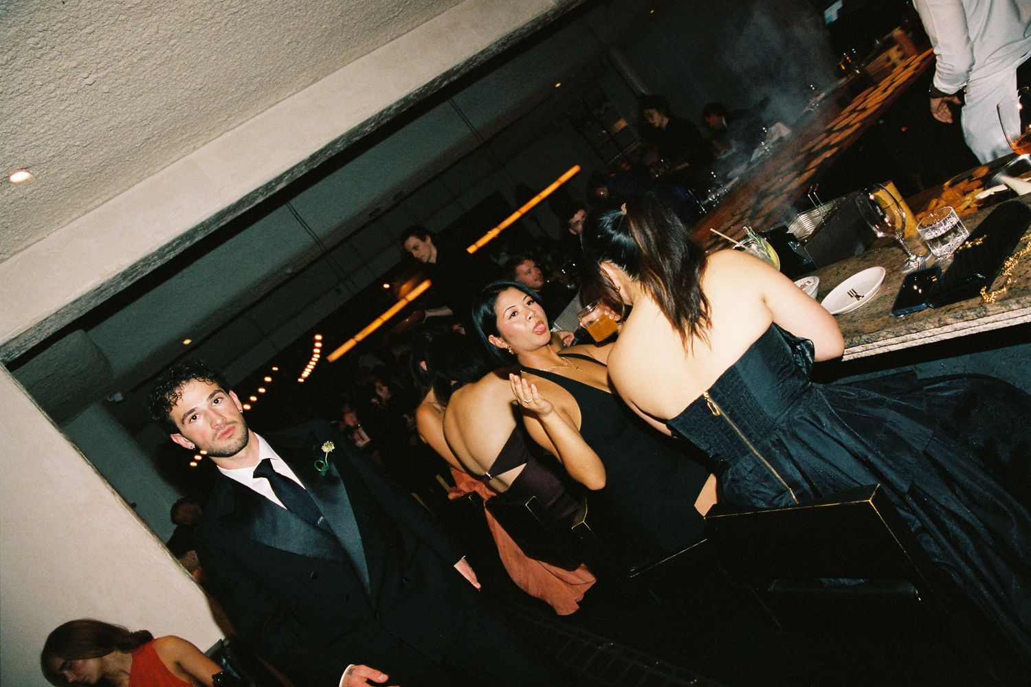 Wedding reception guests dancing and socializing at dimly lit venue bar area in formal attire.