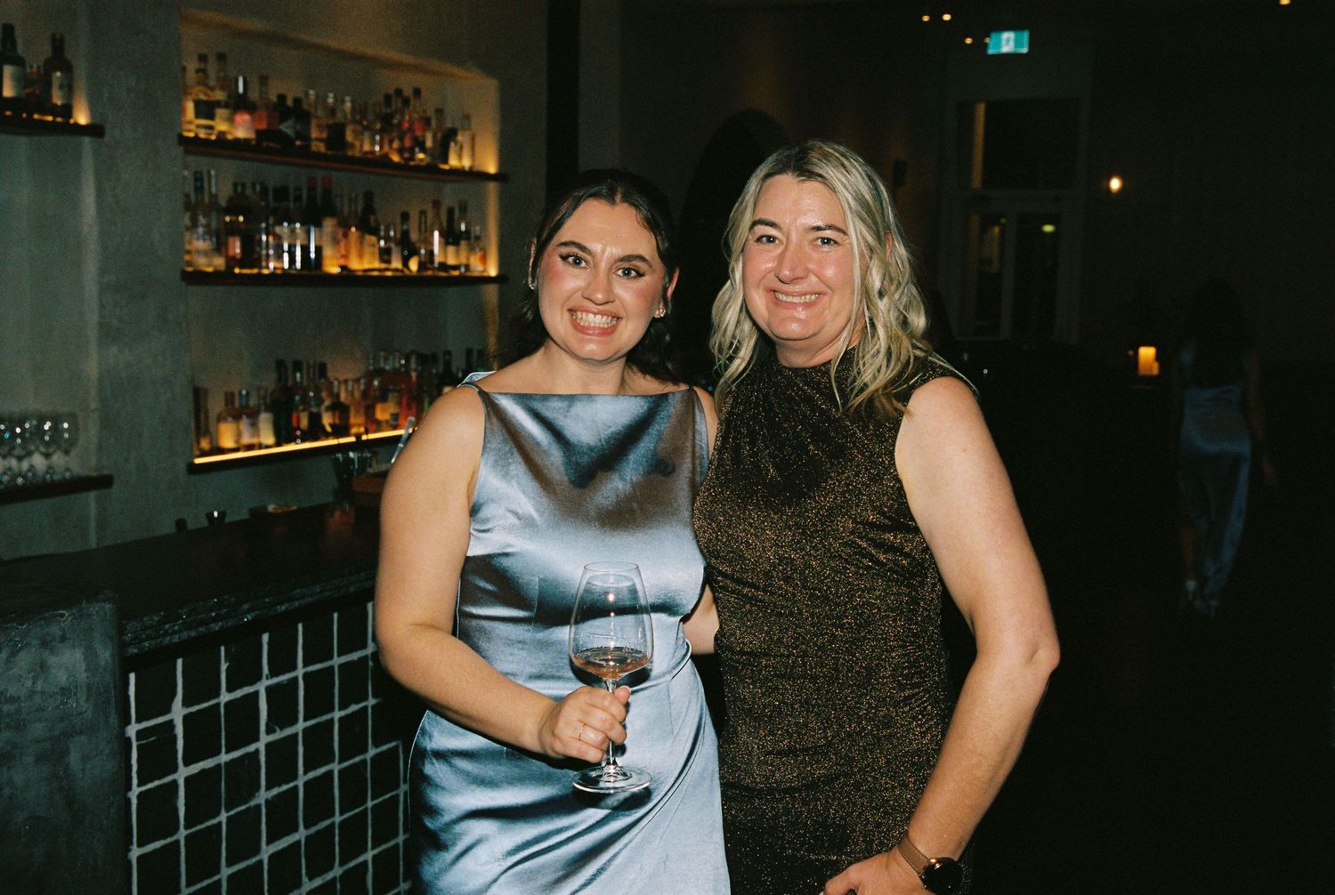 Two women smiling together at a wedding reception bar, one holding a wine glass.