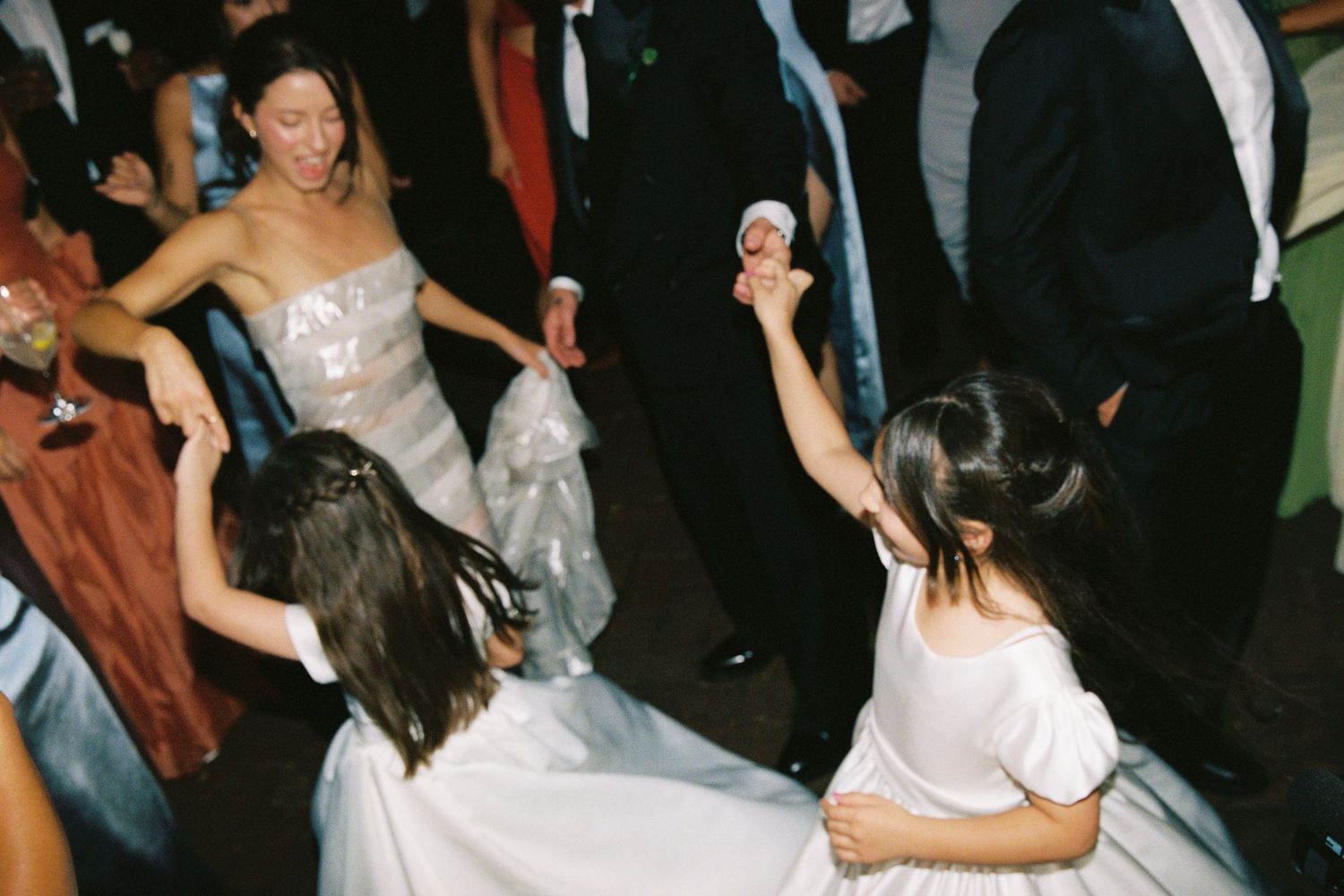 Bride in silver gown dancing with young flower girls at a wedding reception.