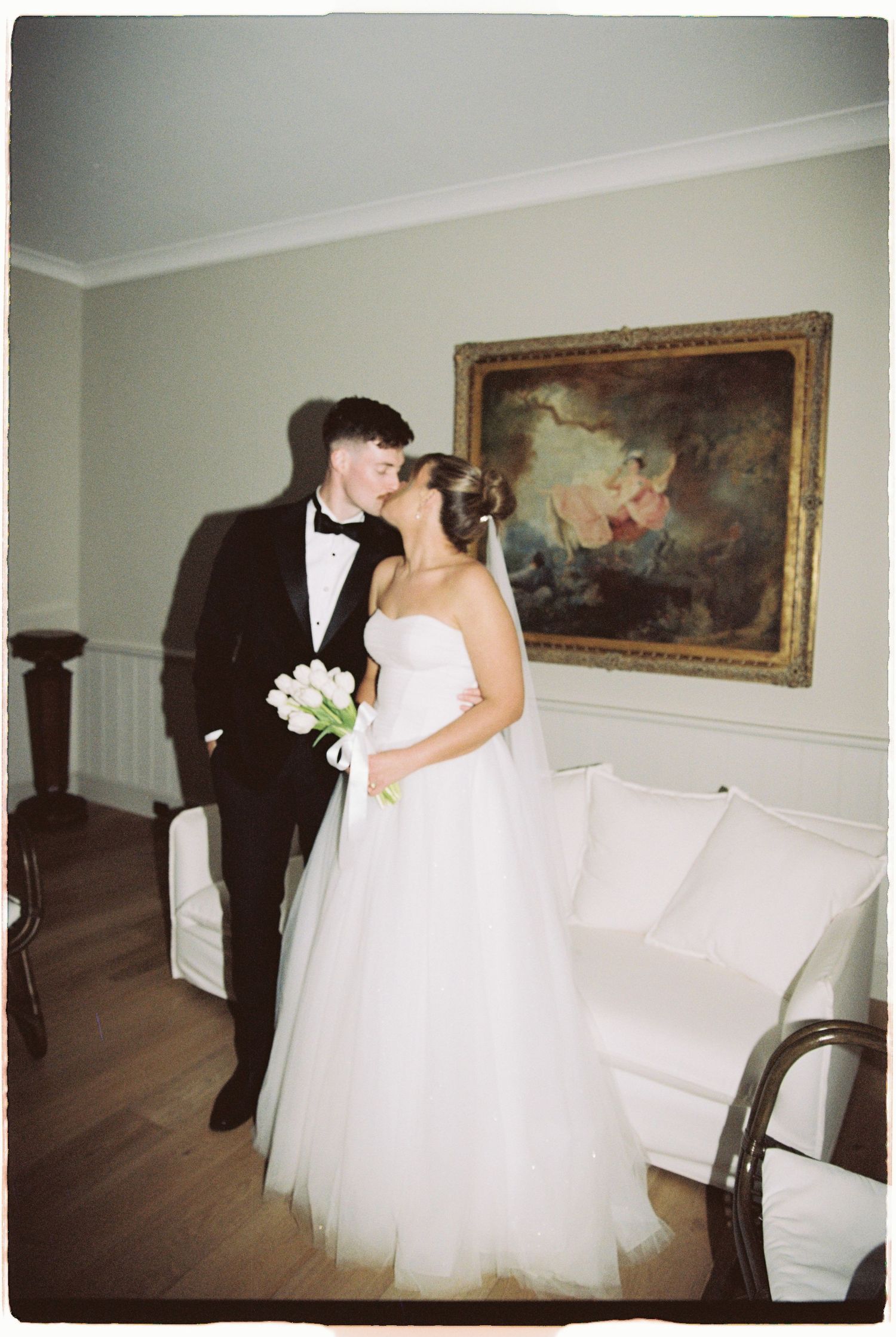 Bride and groom sharing a kiss indoors beside ornate painting, bride holding white tulip bouquet.