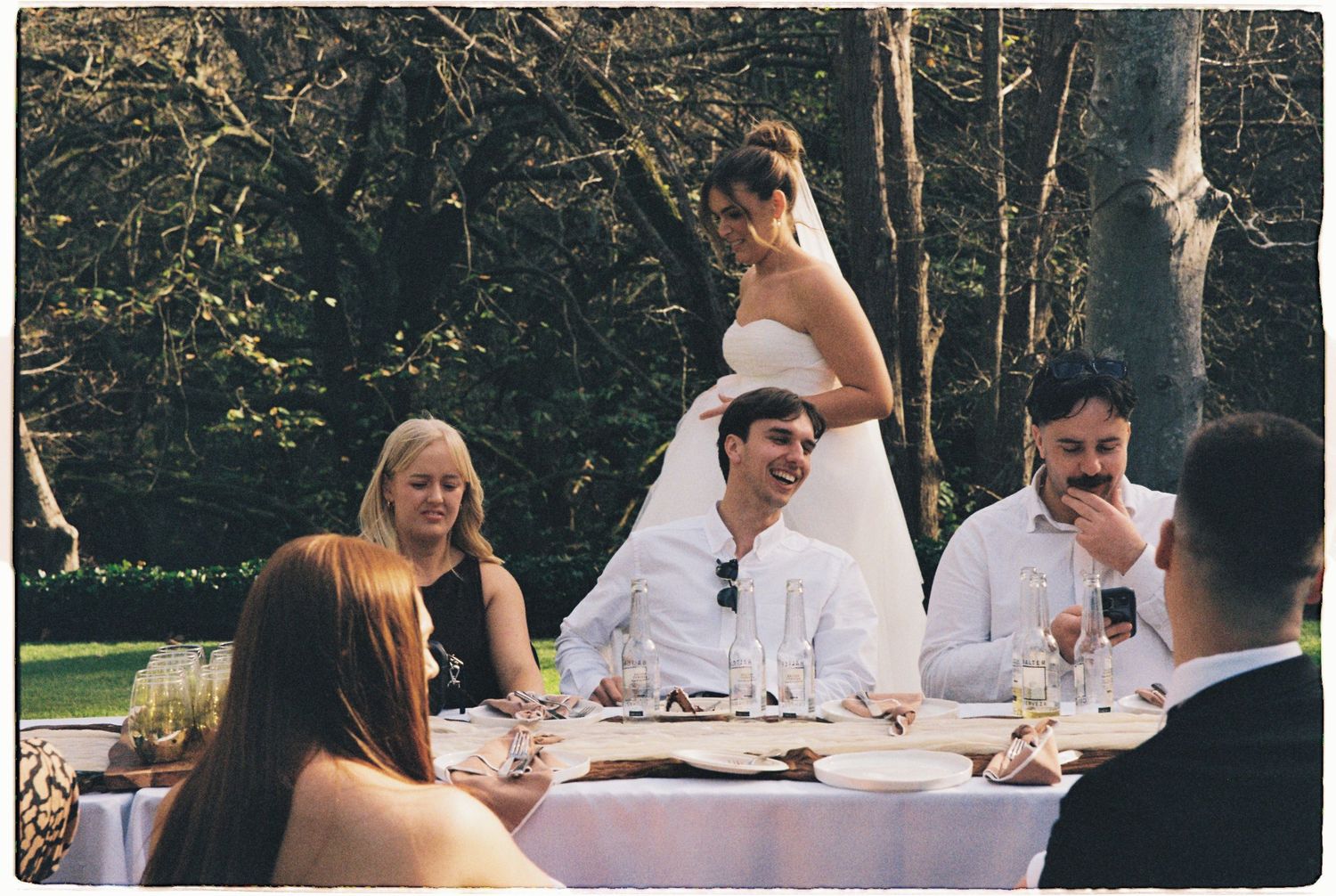 Bride stands behind groom at outdoor wedding reception table with smiling guests seated in a sunlit garden.