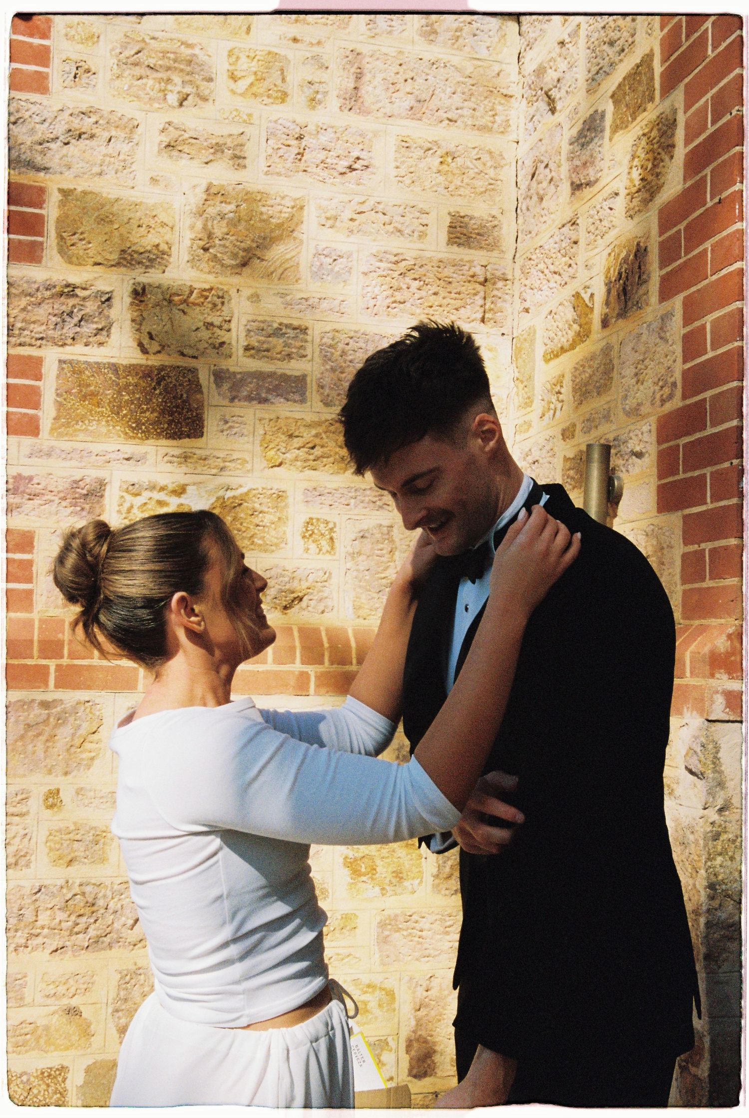 Bride and groom share a romantic moment against a rustic sandstone brick wall, she adjusts his bow tie.