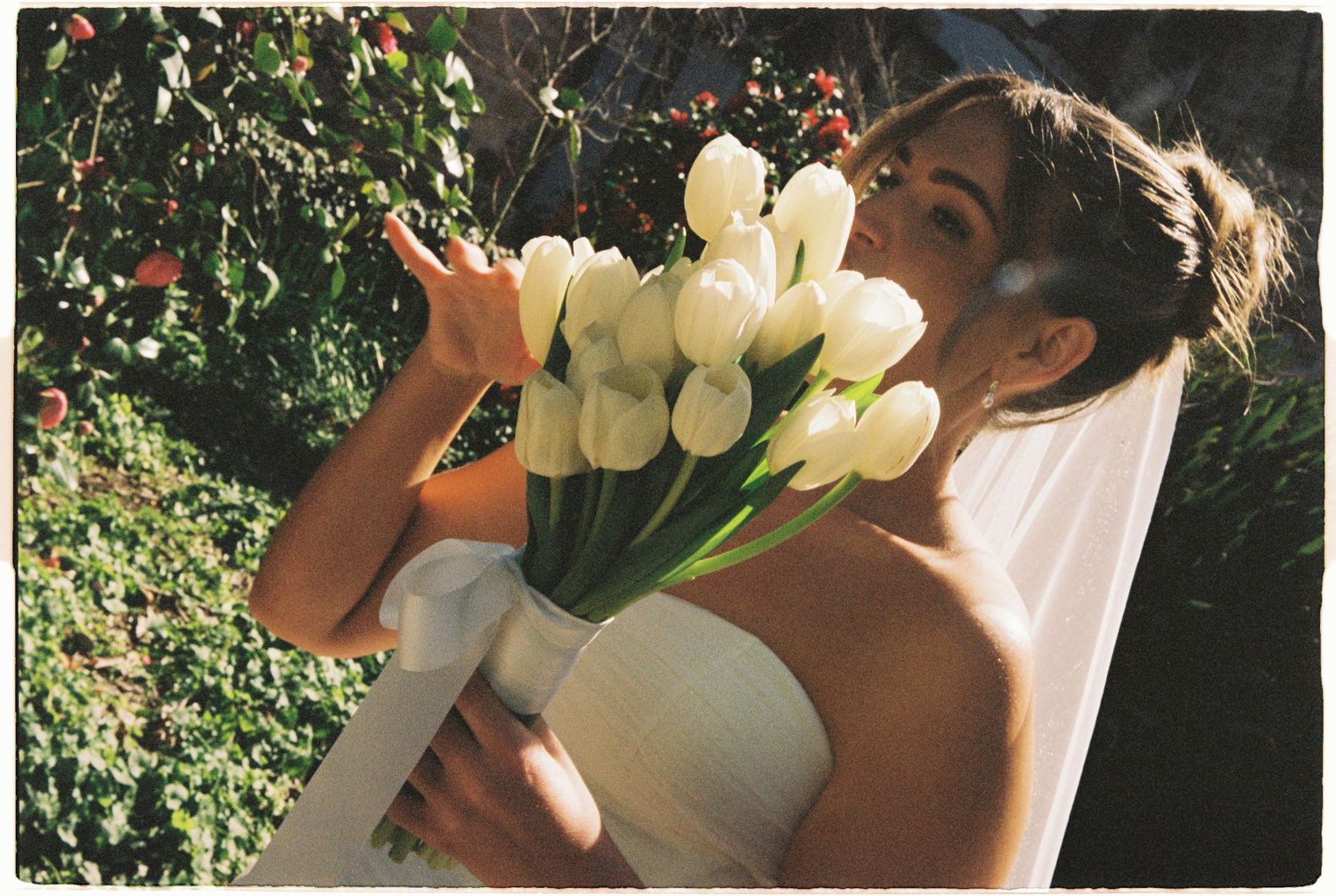 Bride in white veil holding large bouquet of white tulips in lush garden setting.
