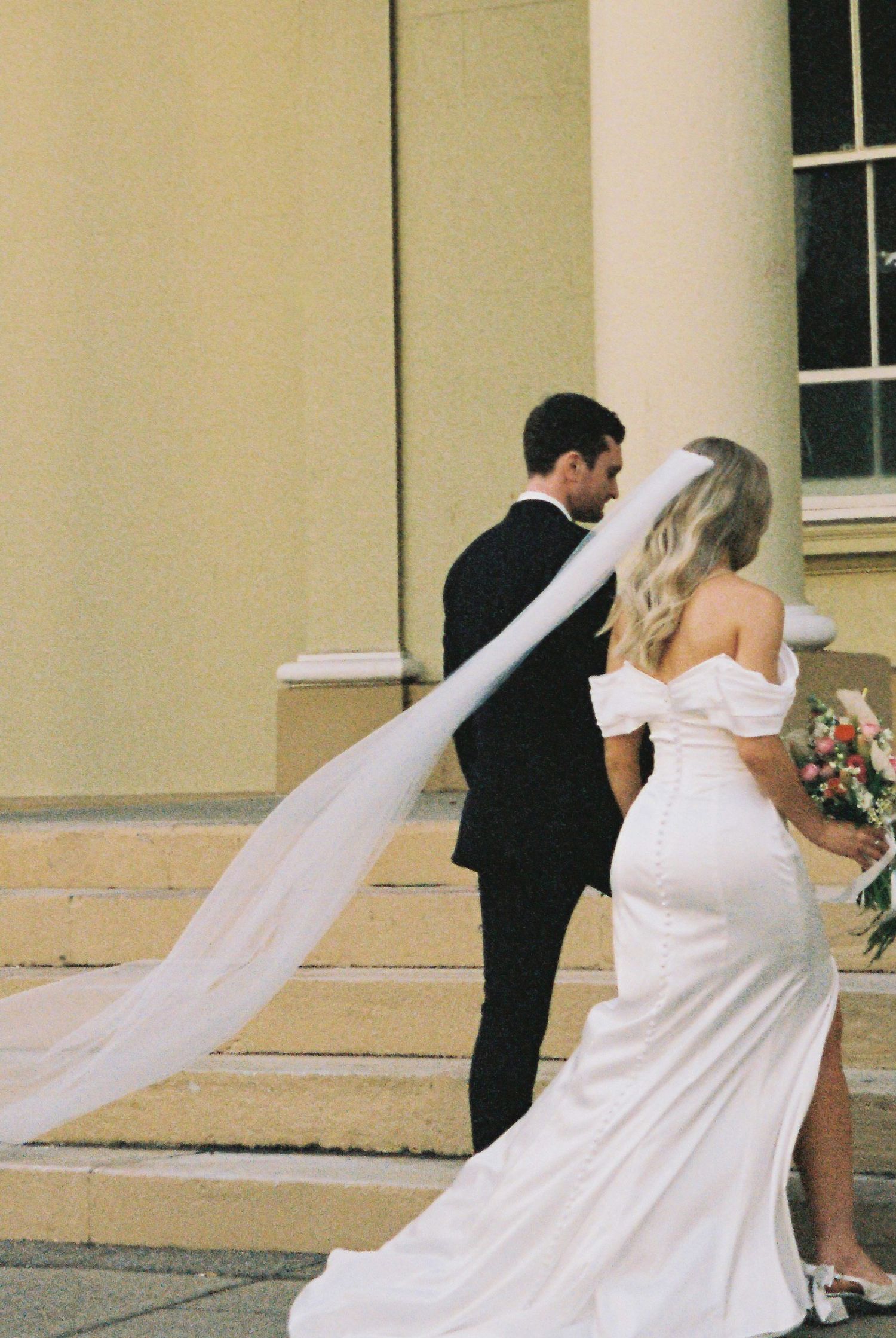 Bride's cathedral veil flows in wind as couple walks up stone steps outside elegant cream-colored venue.
