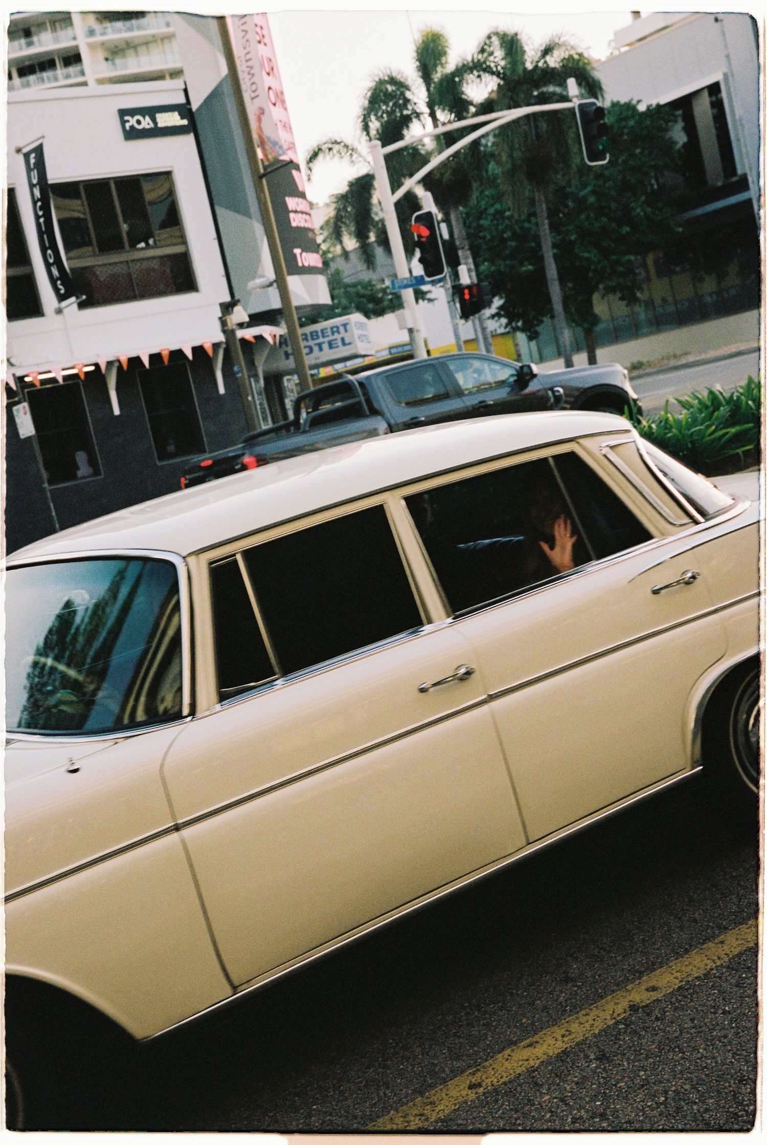 Vintage cream Mercedes-Benz wedding car drives through city street with passenger waving from window.