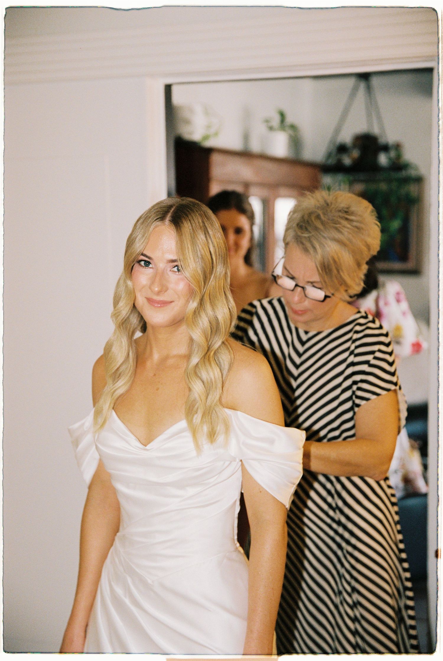 Bride in white off-shoulder gown getting dressed with help from older woman in striped dress, mirror reflection in background.
