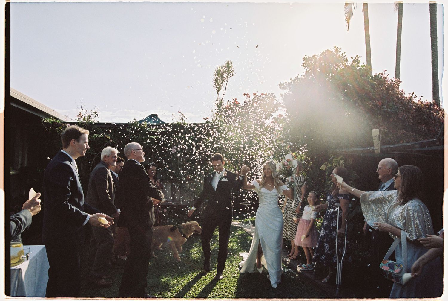 Newlyweds celebrate with confetti in a sunlit garden surrounded by wedding guests.
