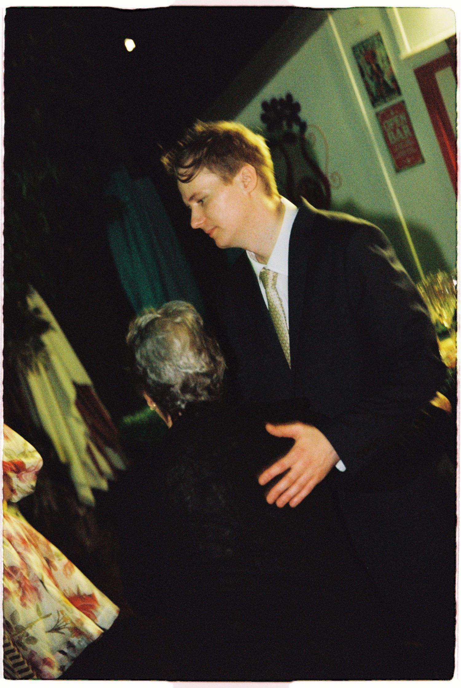 A man in a black suit and tie leans forward speaking to an elderly guest at a wedding reception.