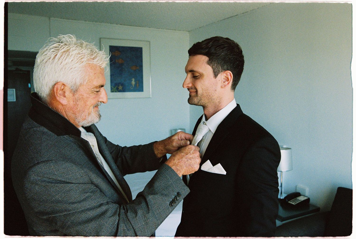 Silver-haired father adjusting his sons tie as groom prepares for wedding in a hotel room.