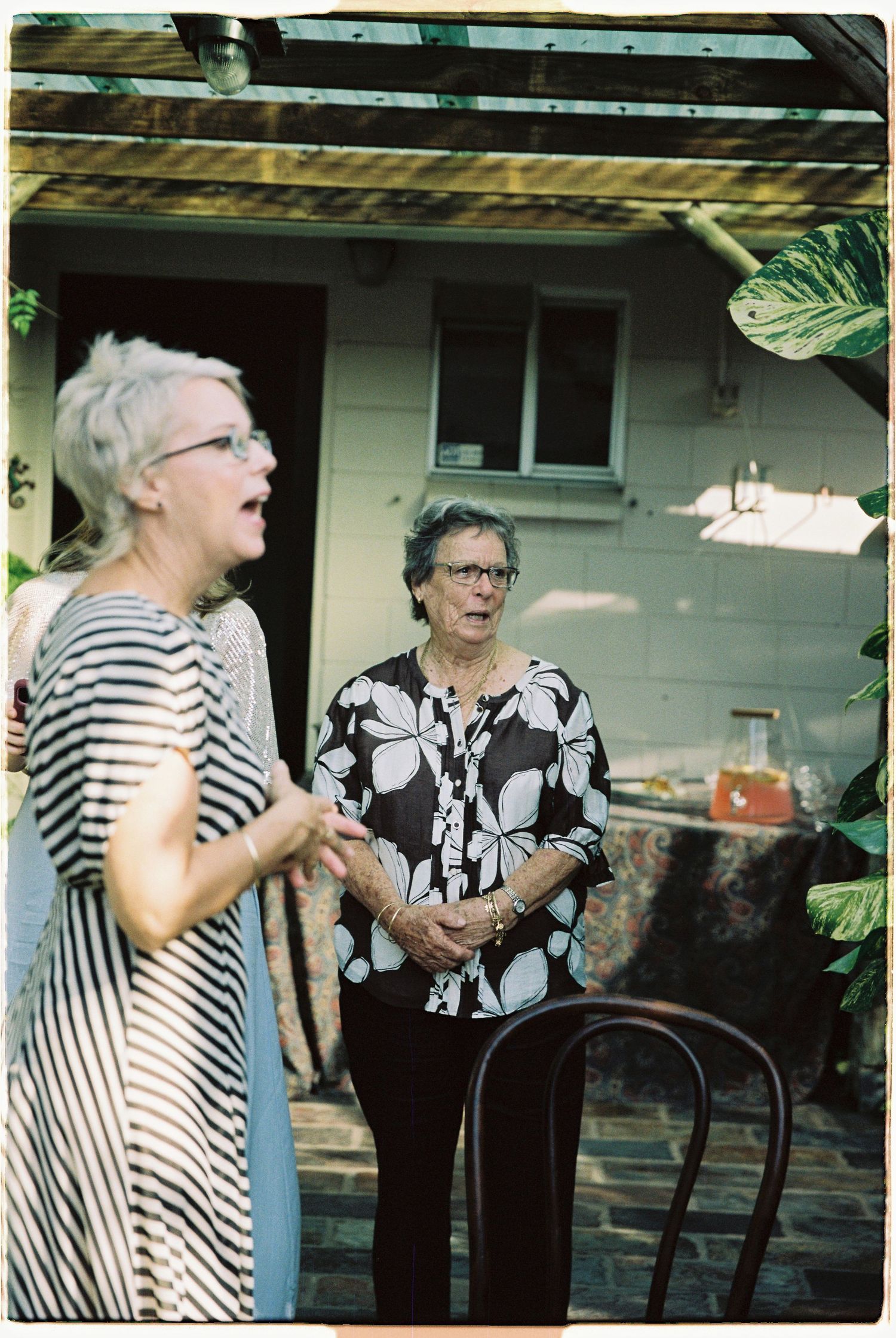 Two older women in casual attire converse on outdoor patio surrounded by tropical plants at wedding.