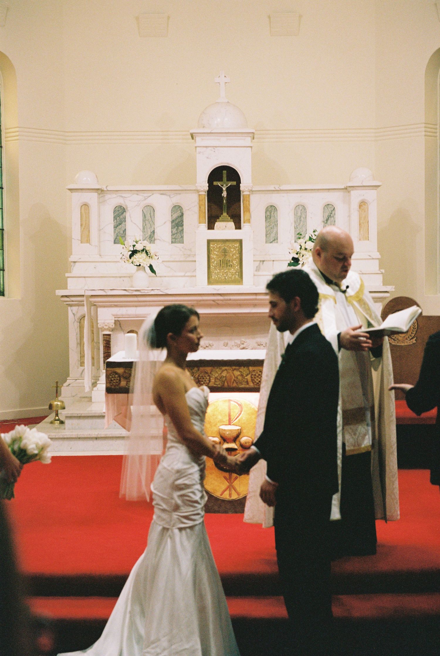 Bride and groom hold hands during Catholic wedding ceremony before ornate white altar.
