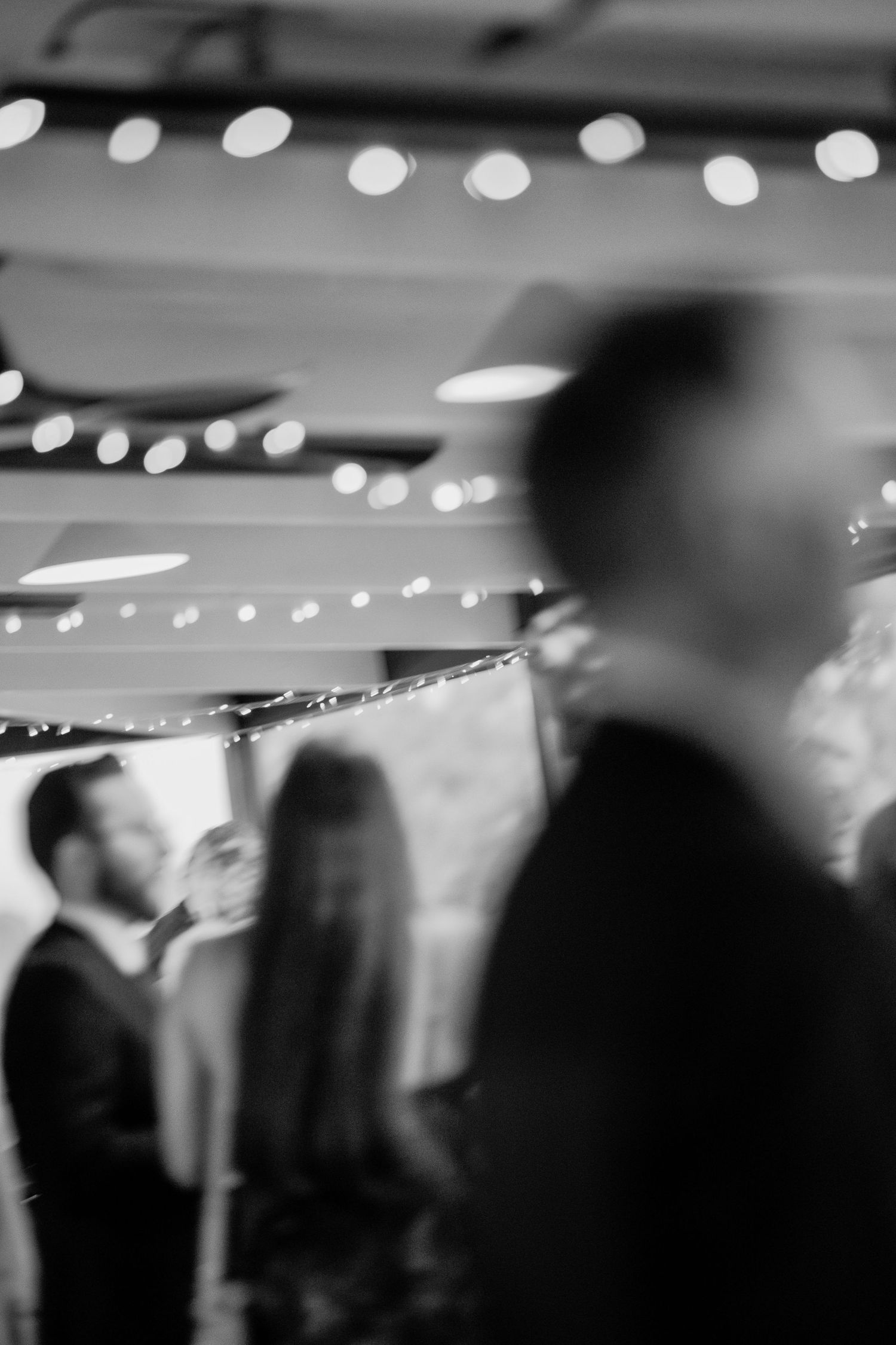 Blurry black and white photo of elegantly dressed guests mingling at an indoor event with bokeh string lights.