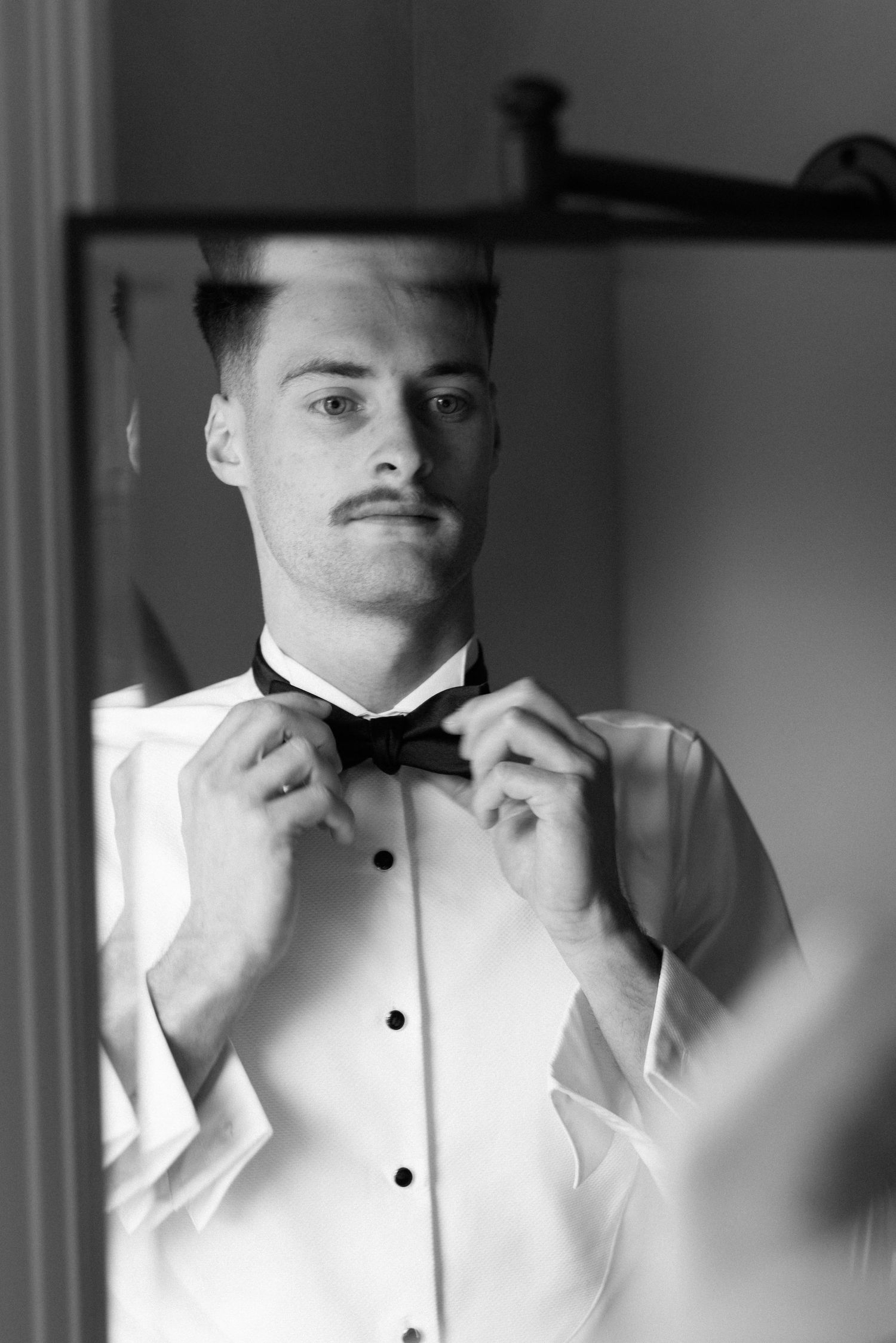 Groom adjusting bow tie in mirror reflection, wearing white tuxedo shirt, black and white wedding photo.