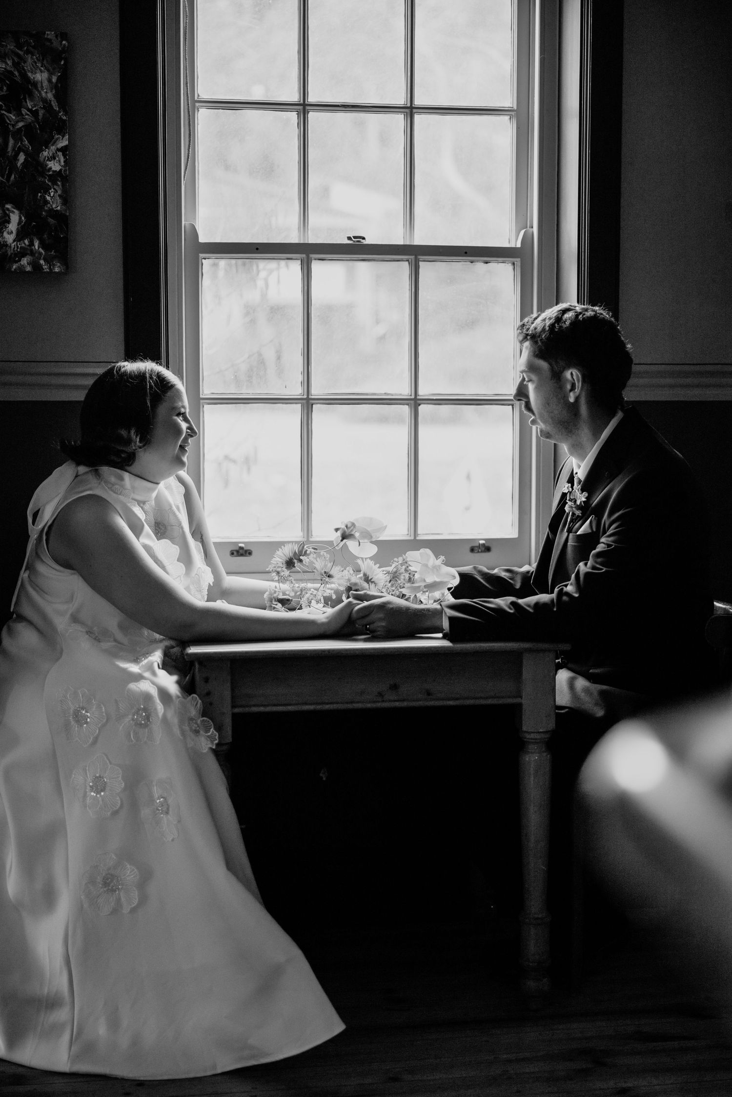 Black and white photo of a bride and groom holding hands across a table by a window on their wedding day.