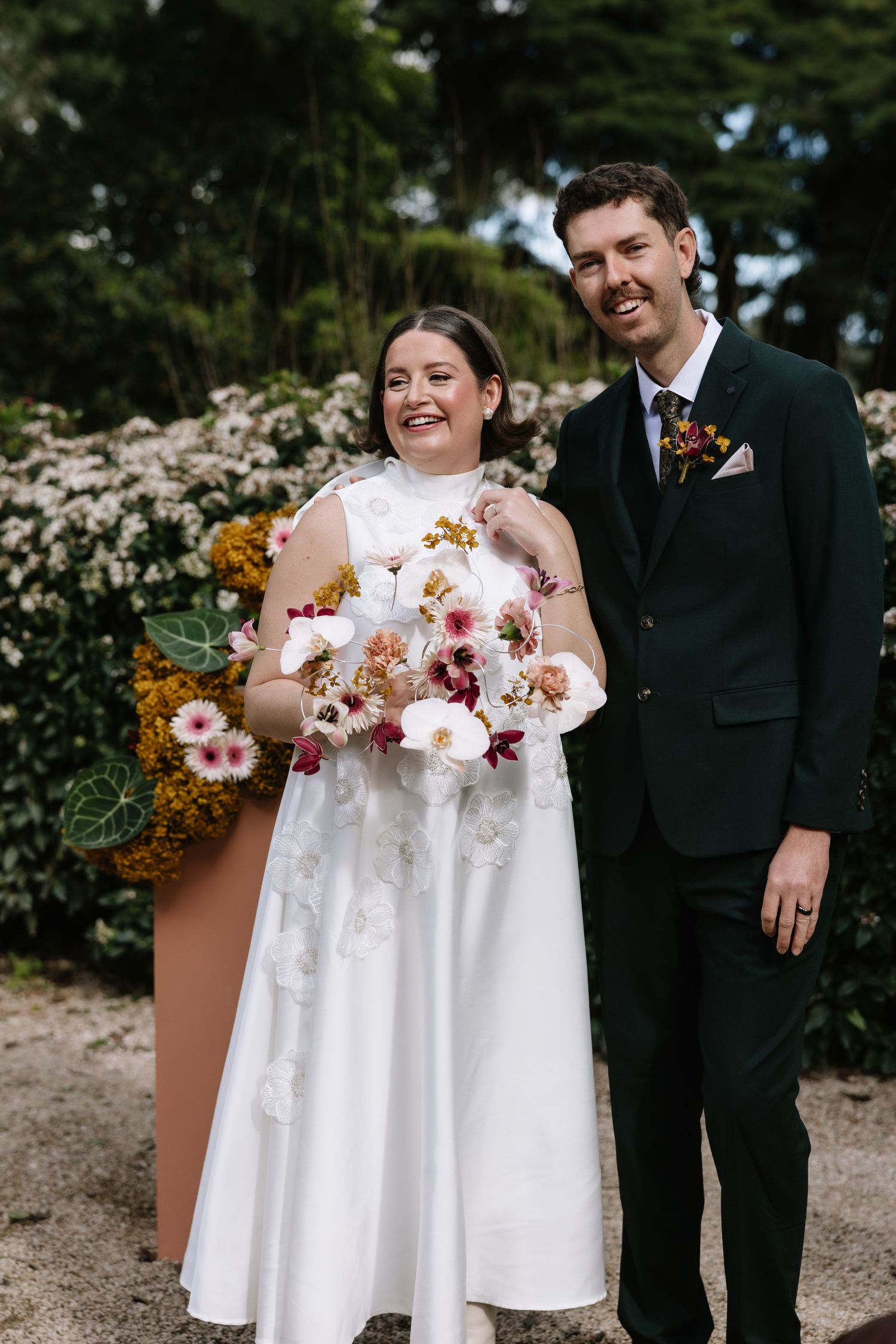 Bride in white gown holding pink floral bouquet stands with groom in dark suit outdoors at garden wedding.