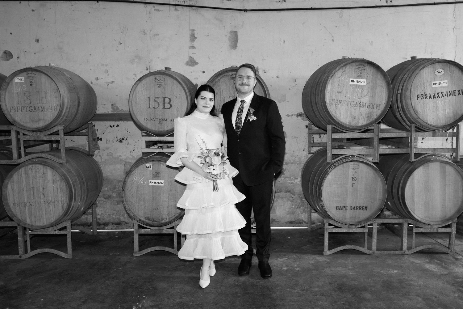 Bride and groom posing together in front of wine barrels in rustic winery cellar.