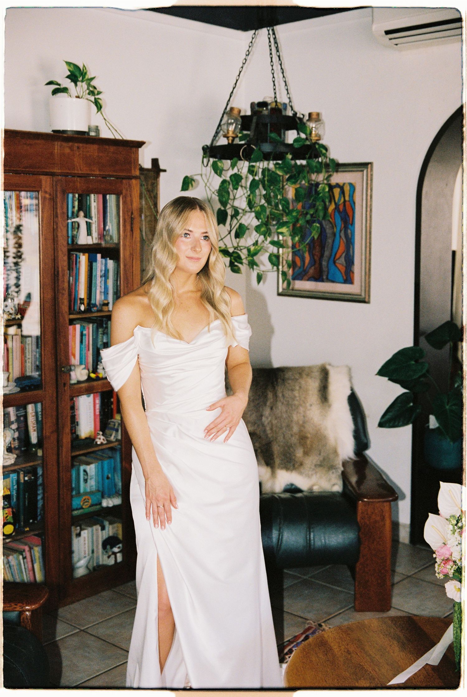 Blonde bride in off-shoulder white wedding gown posing in a cozy home interior with bookshelves.