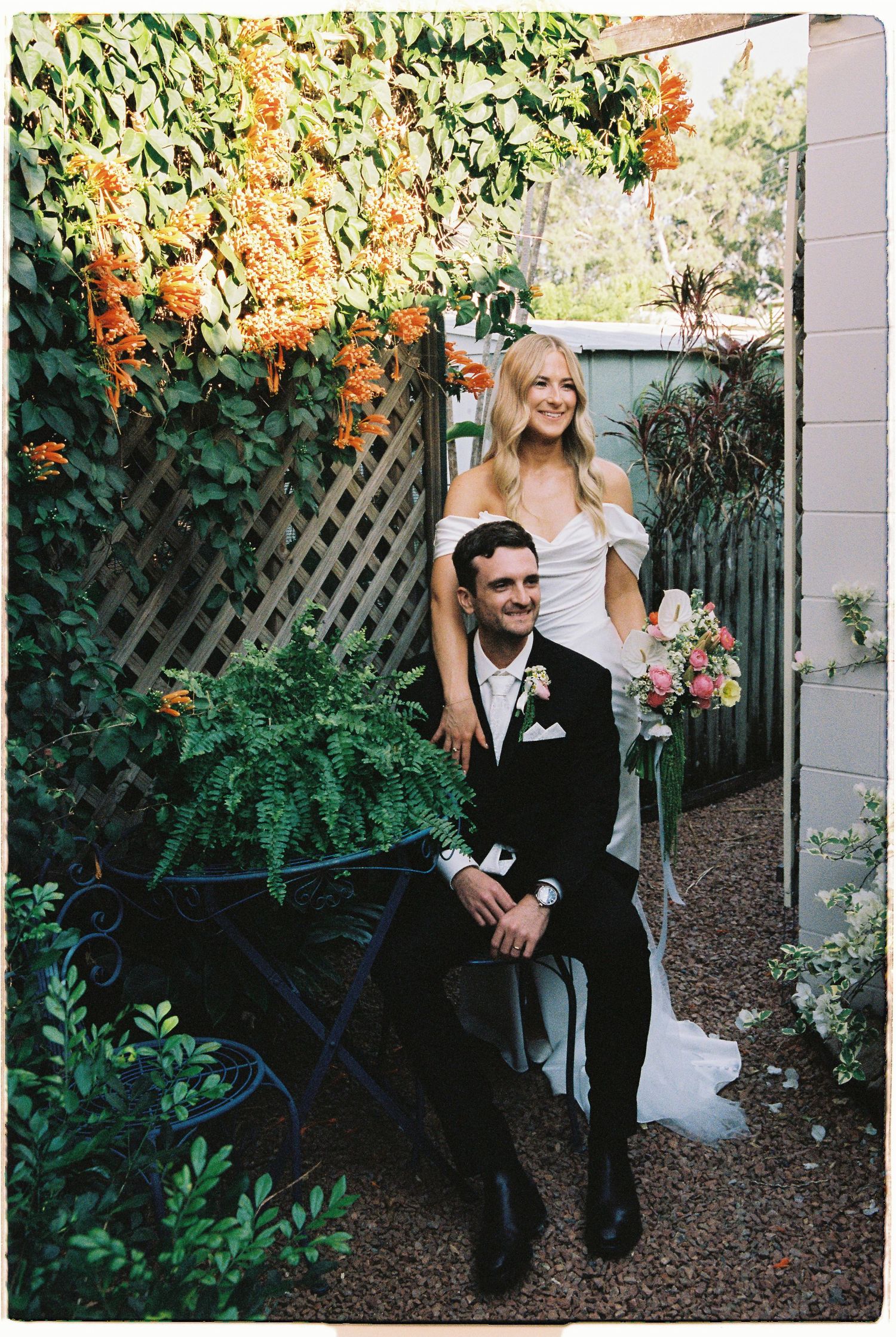 Bride and groom posing in lush garden with orange flowering vines at wedding.