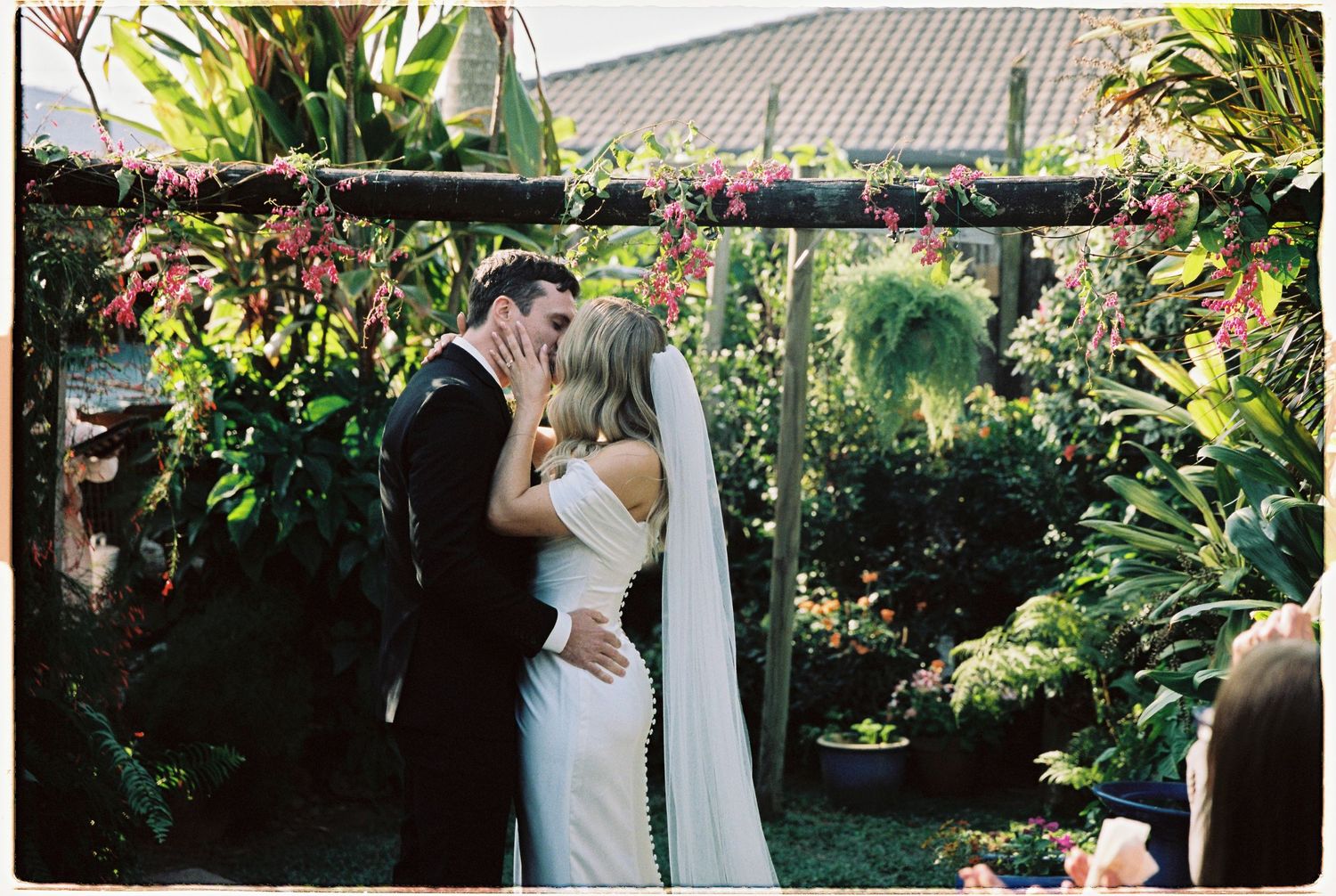 Bride and groom sharing first kiss under floral arch in tropical garden ceremony.