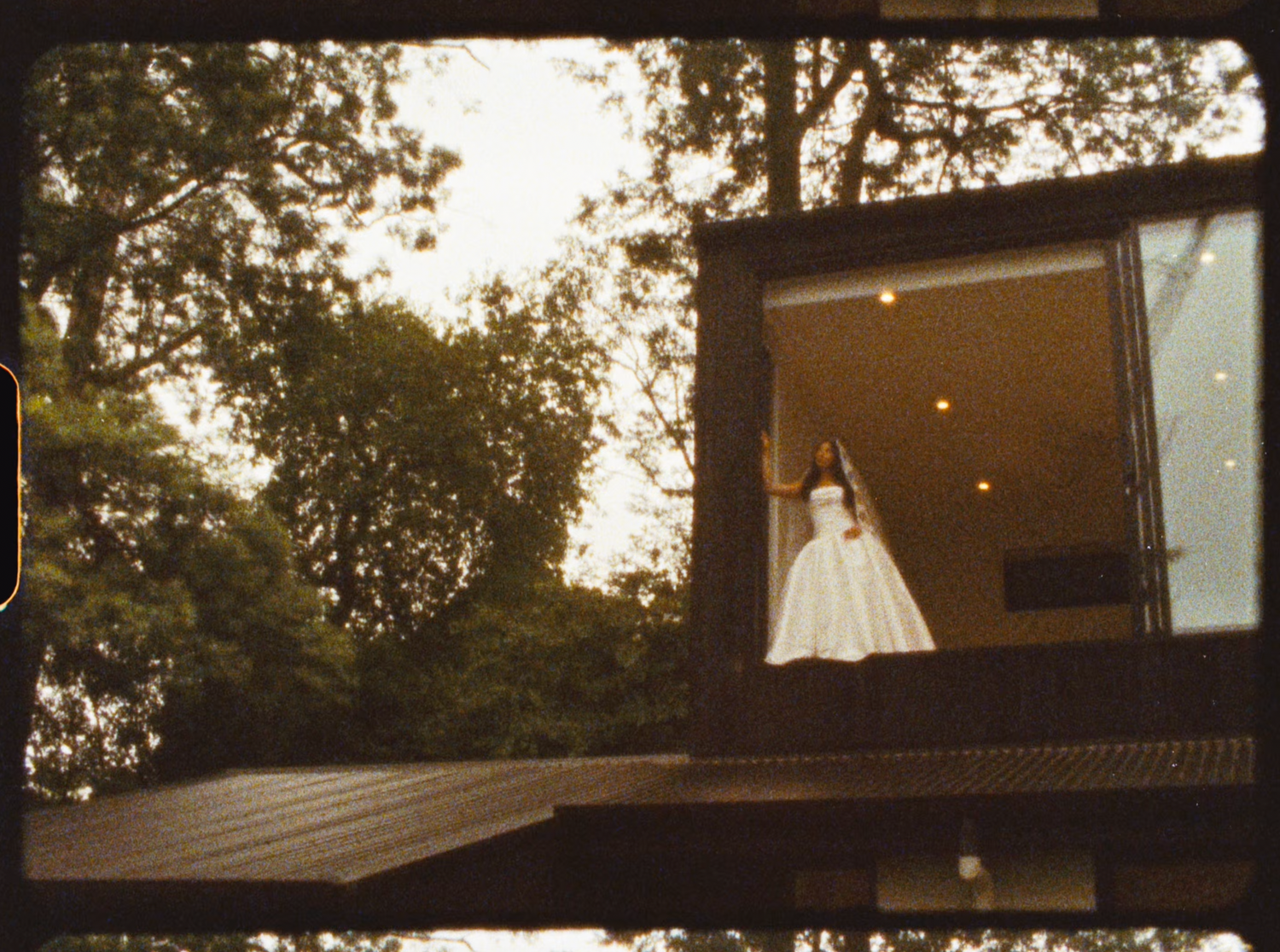 Bride in a white ball gown stands in a modern glass-walled room overlooking lush green trees.