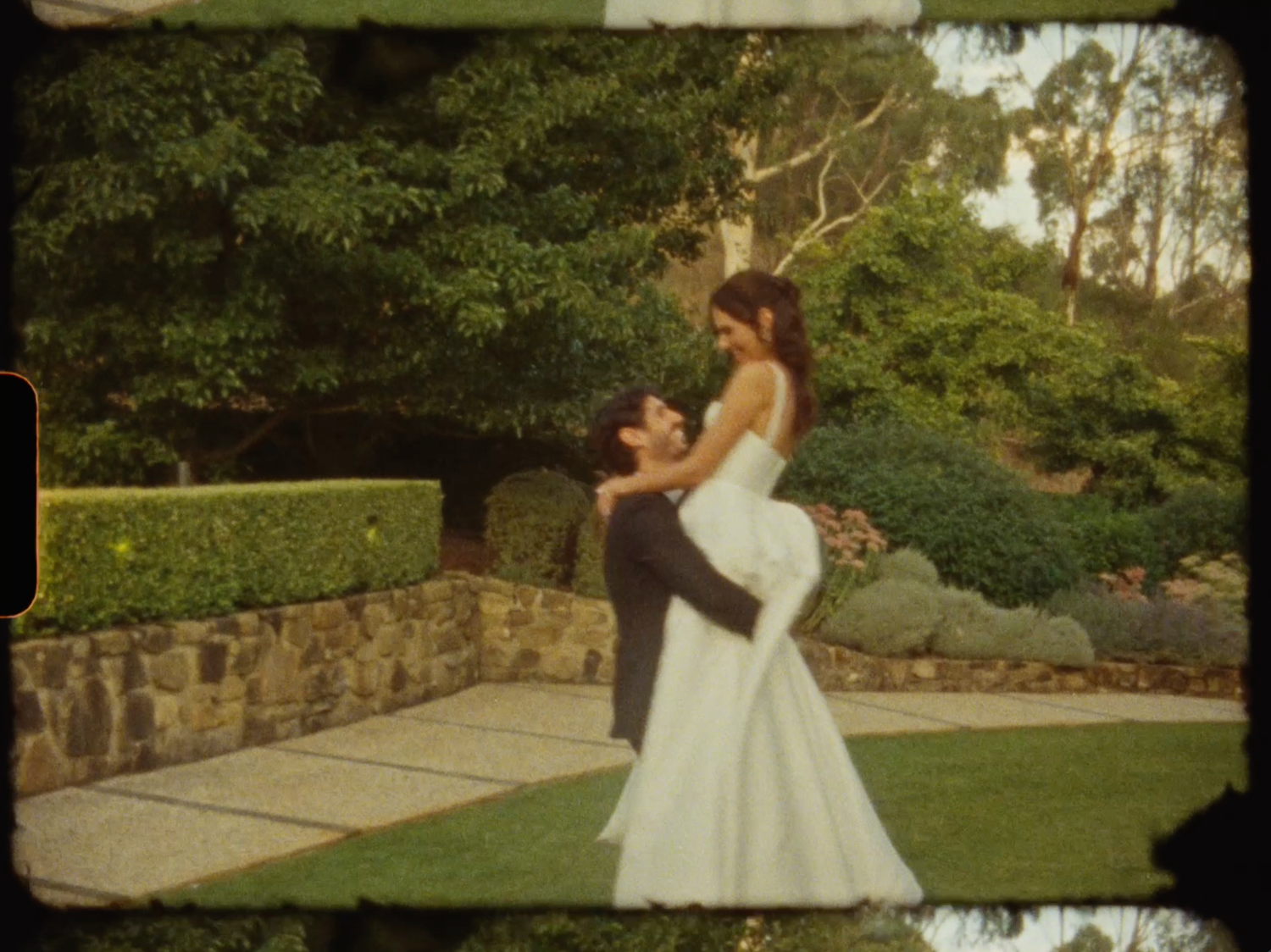 Groom lifts bride in white wedding gown in a romantic garden setting.