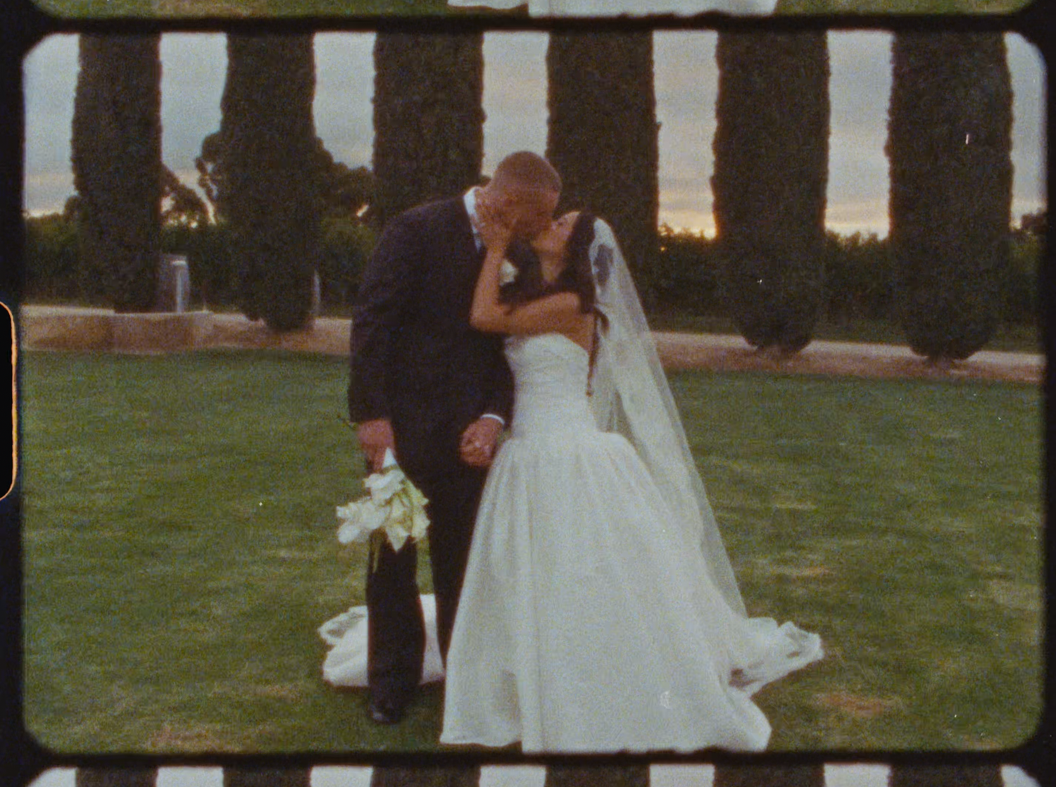 Bride and groom sharing romantic kiss on green lawn with tall cypress trees in background at dusk.