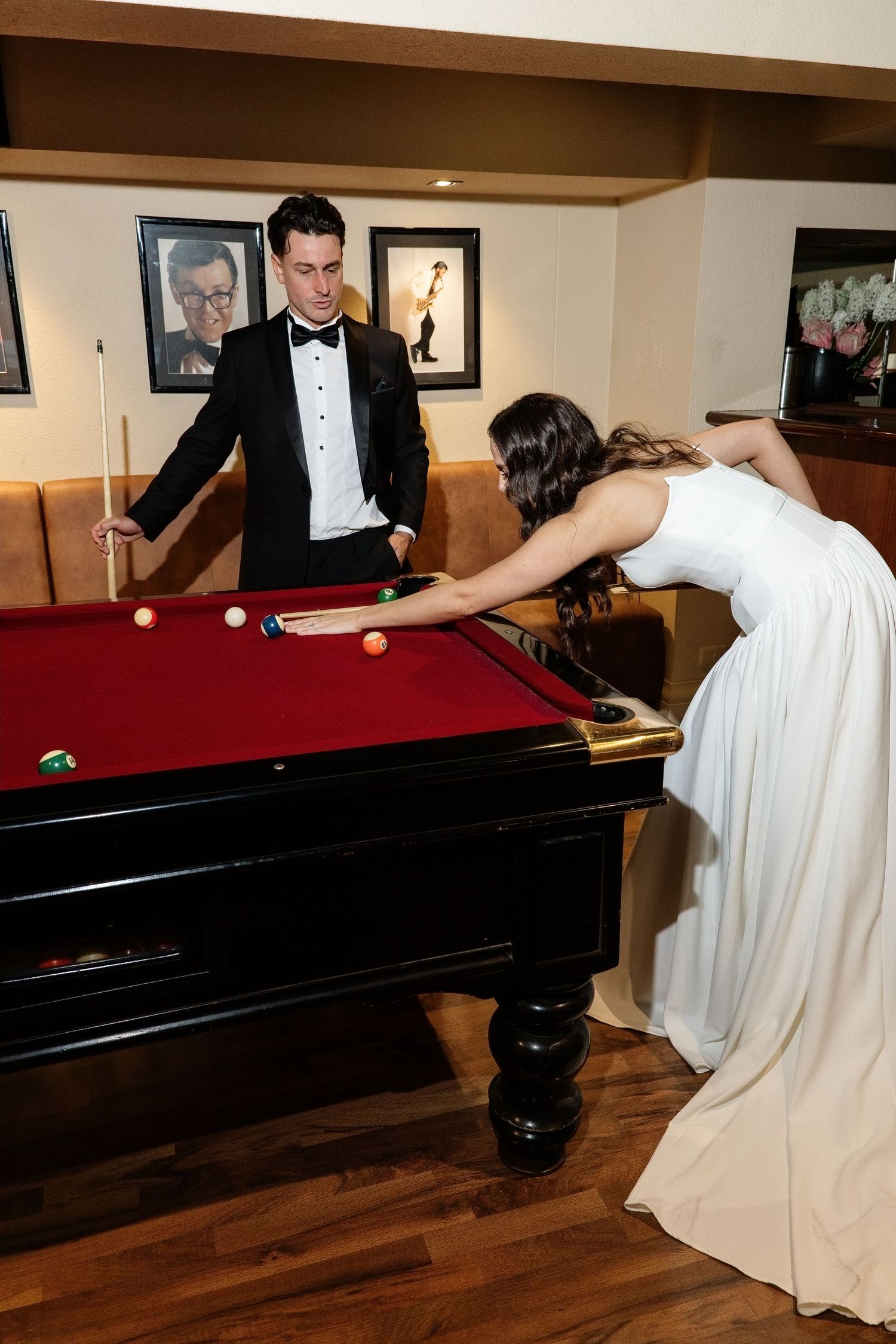 Bride in white gown leaning over red pool table taking a shot while groom in tuxedo watches.