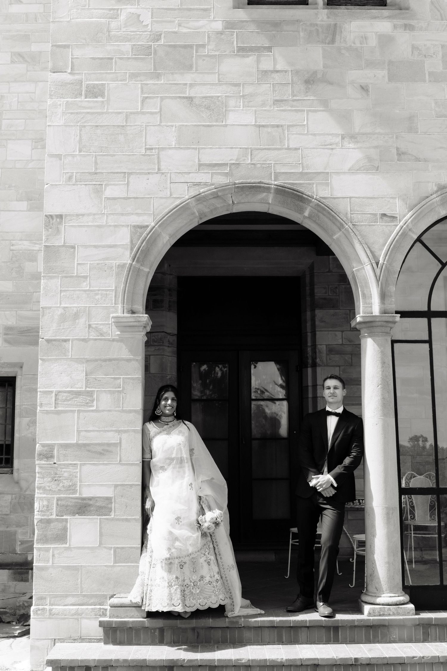 Bride in white lehenga and groom in tuxedo stand beneath stone archway of elegant venue.