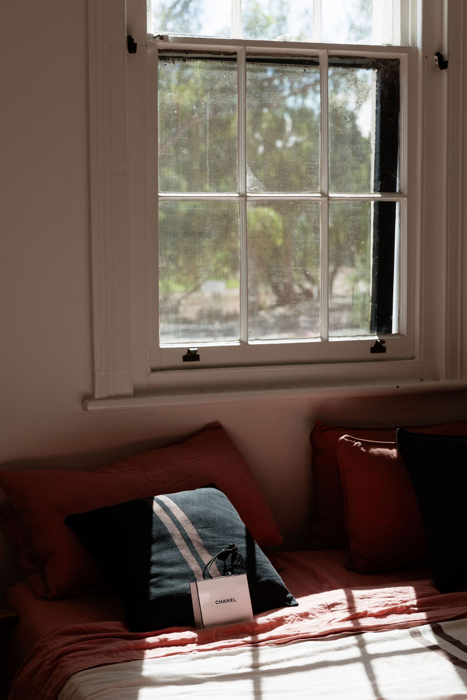 Chanel gift bag on red bedding near sunlit vintage window with garden view.