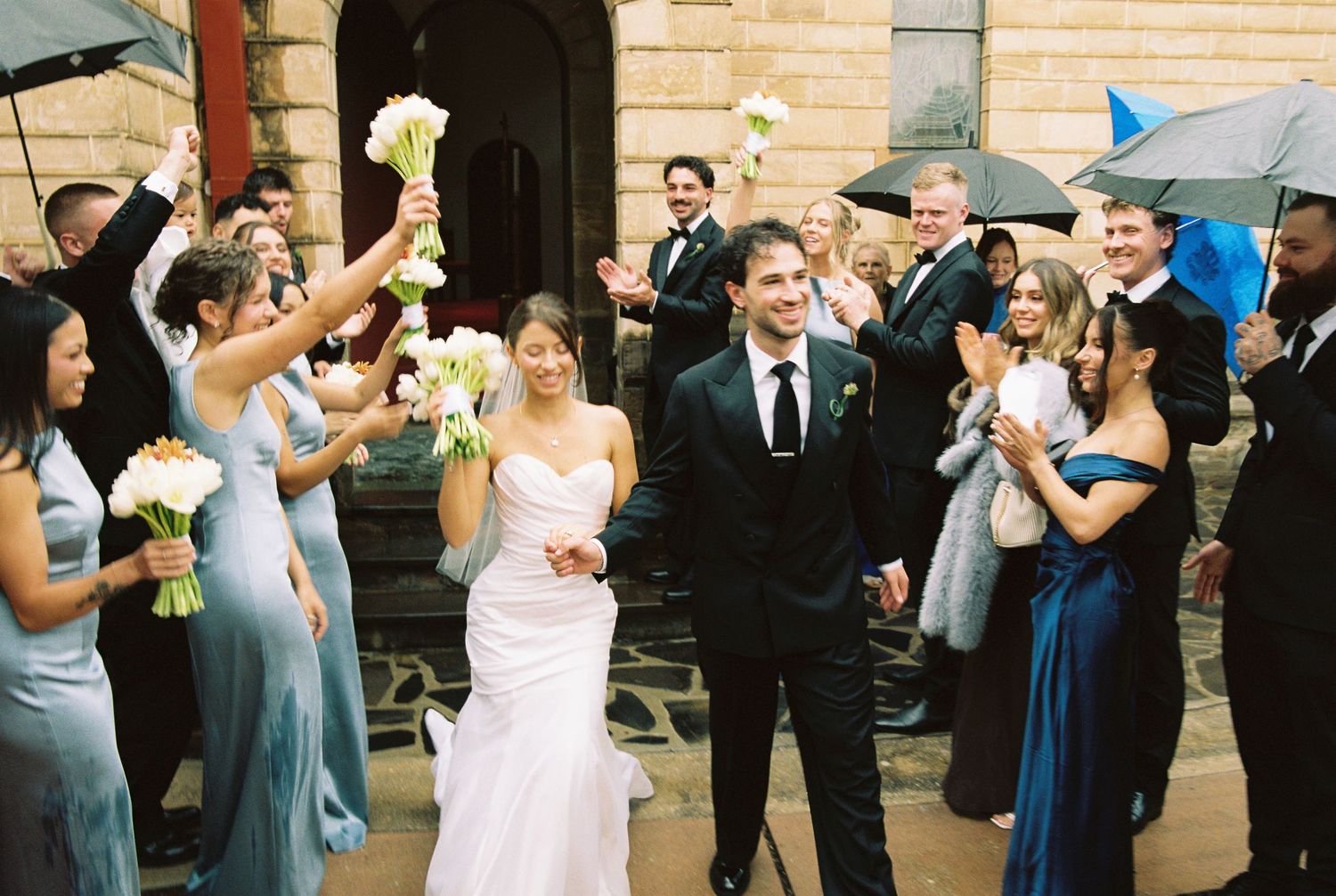 Bride and groom exit church surrounded by cheering wedding party holding bouquets aloft in light rain.