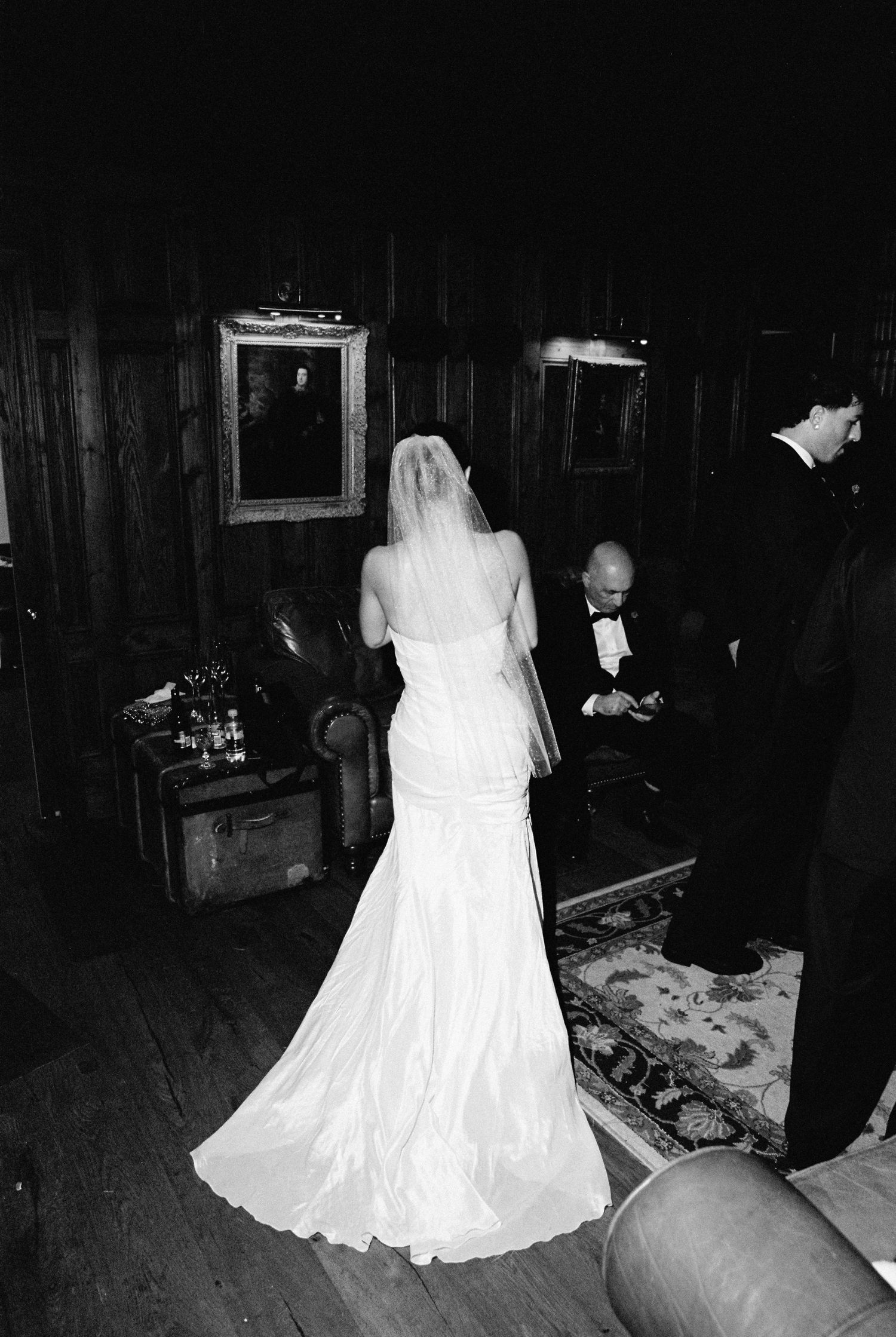 Bride in elegant white gown with veil stands in dark wood-paneled room during wedding.