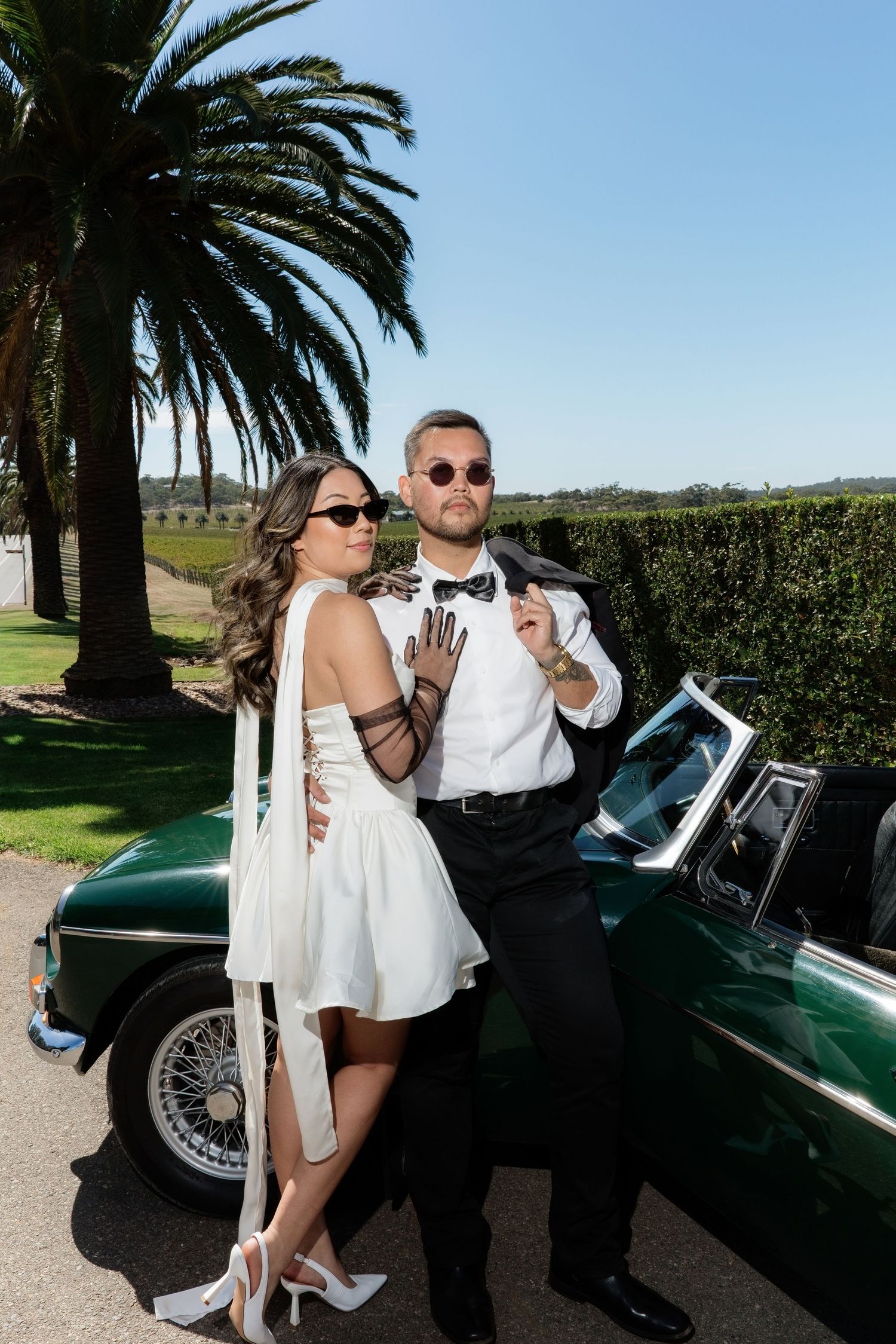 Stylish couple in wedding attire posing by a vintage green convertible car with palm trees in background.