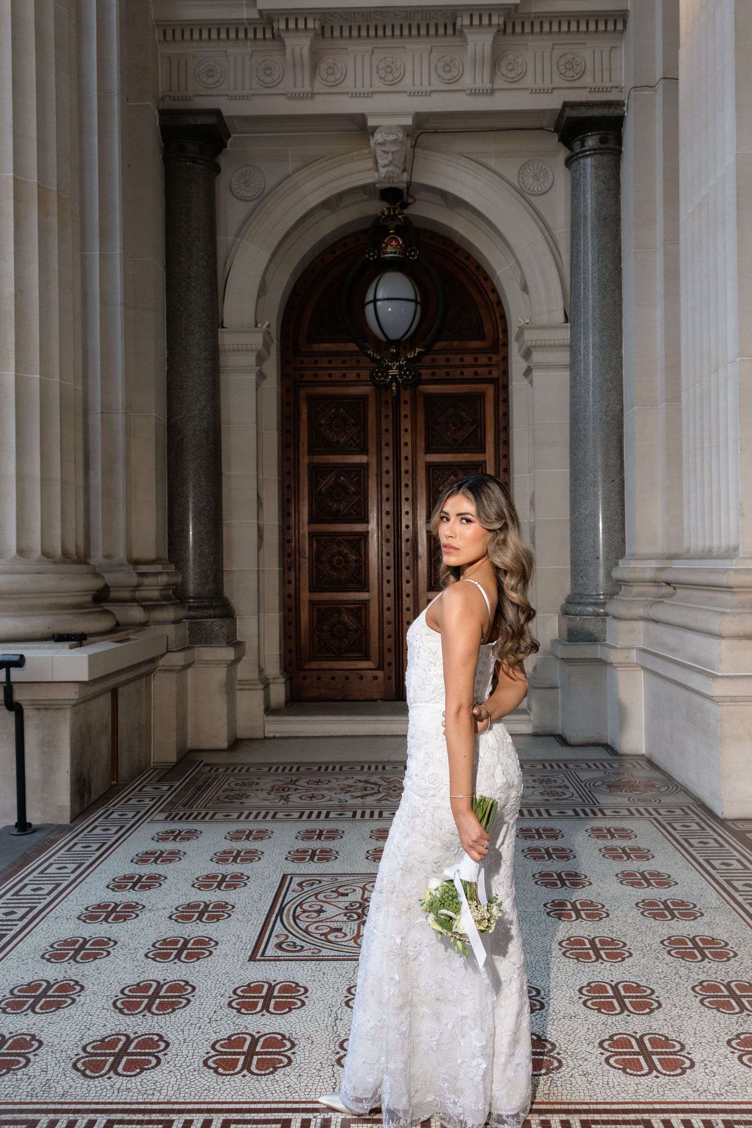 Bride in fitted white lace gown holding bouquet stands on mosaic tiles before ornate wooden doors.
