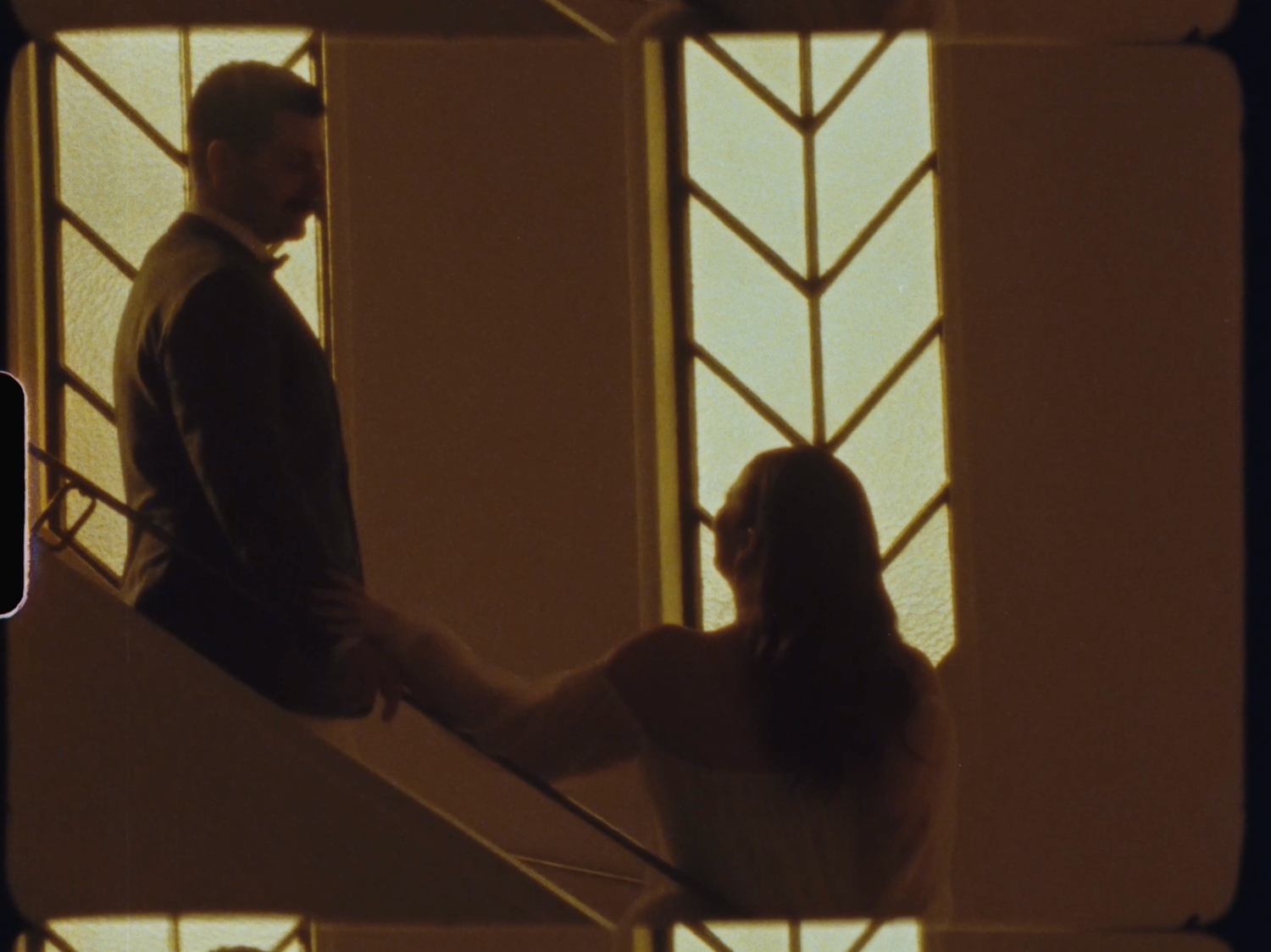 Two silhouetted figures hold hands on a staircase, backlit by ornate Art Deco windows in warm golden light.