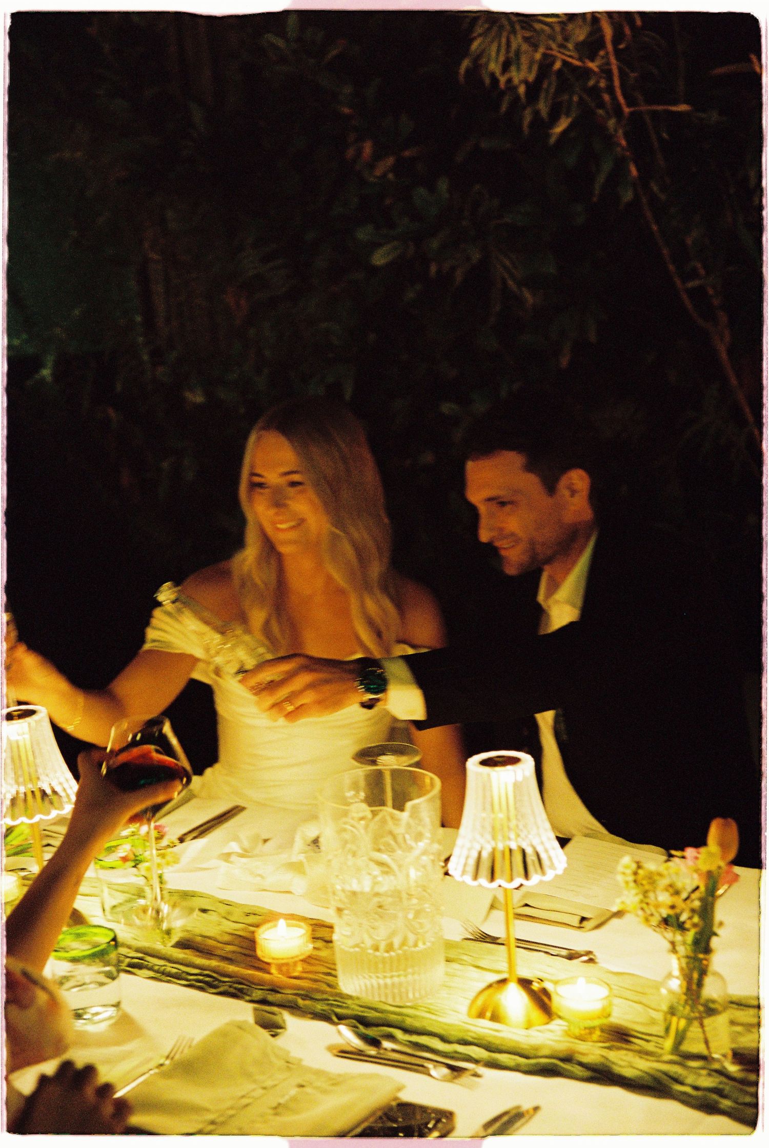 Bride and groom sharing romantic candlelit outdoor wedding reception dinner at night.